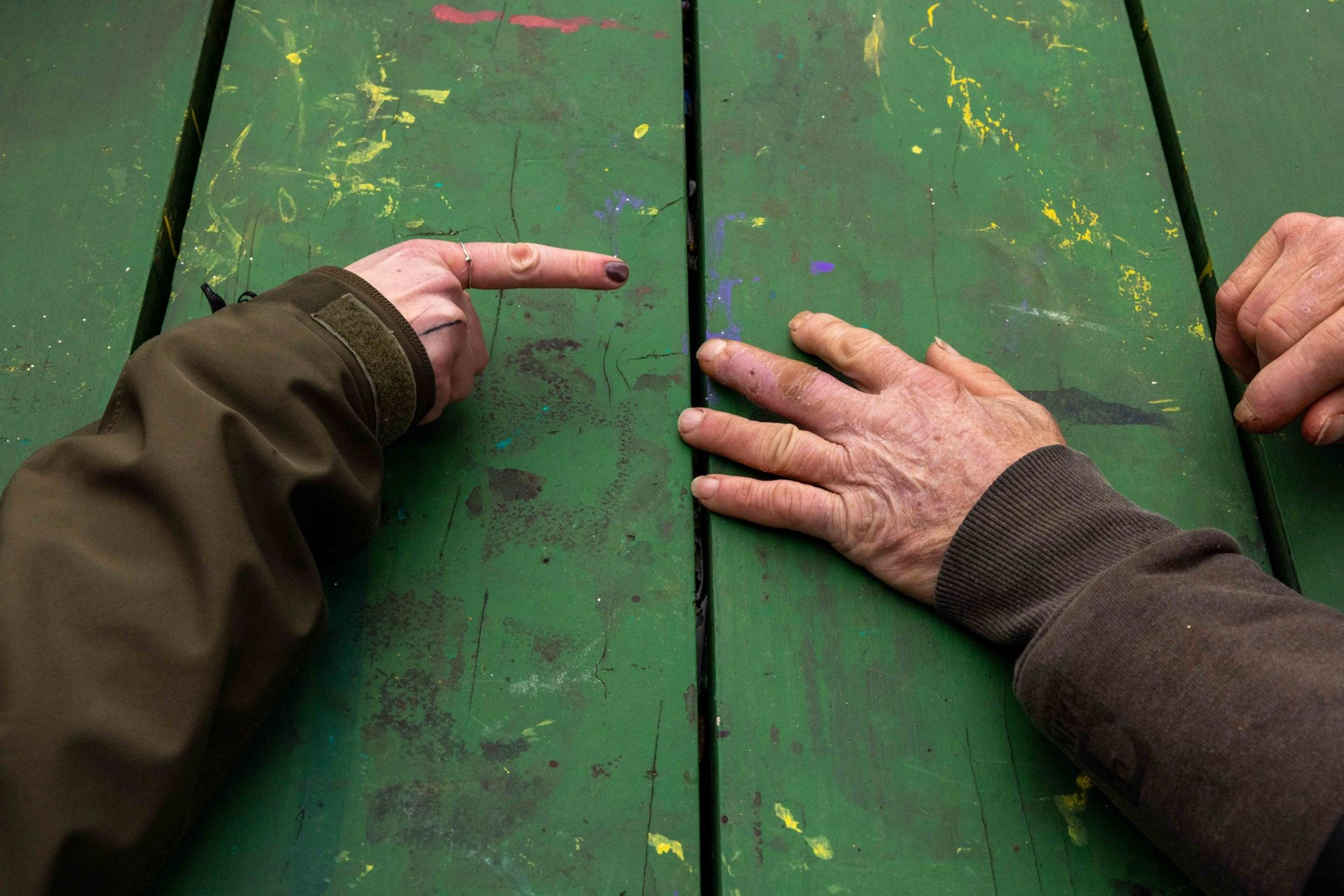 Two hands rest on a weathered, paint-spattered green table. One hand points, while the other is flat, showing different textures and colors.
