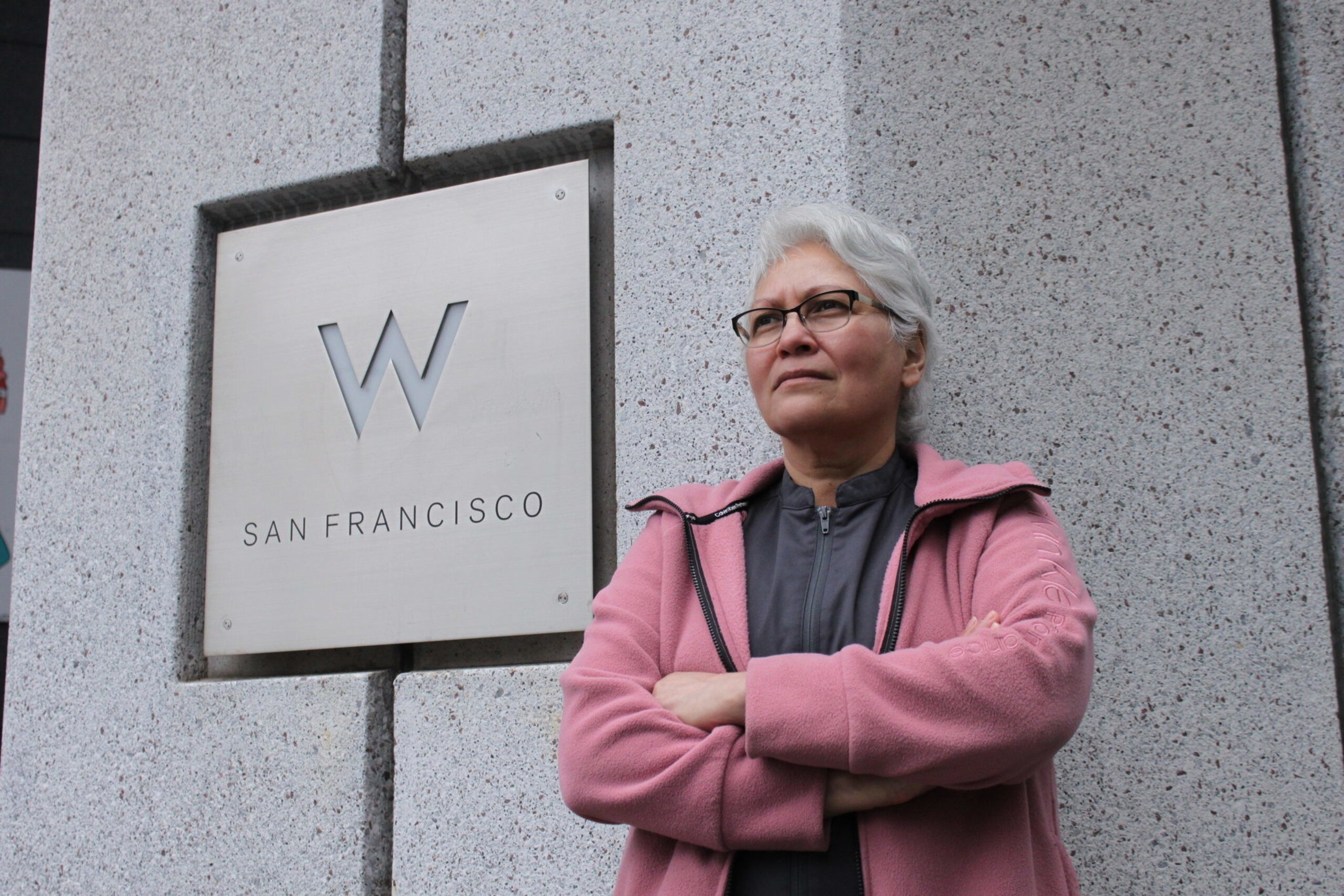 A person with short white hair and glasses stands with arms crossed, wearing a pink jacket, in front of a "W San Francisco" sign on a stone wall.