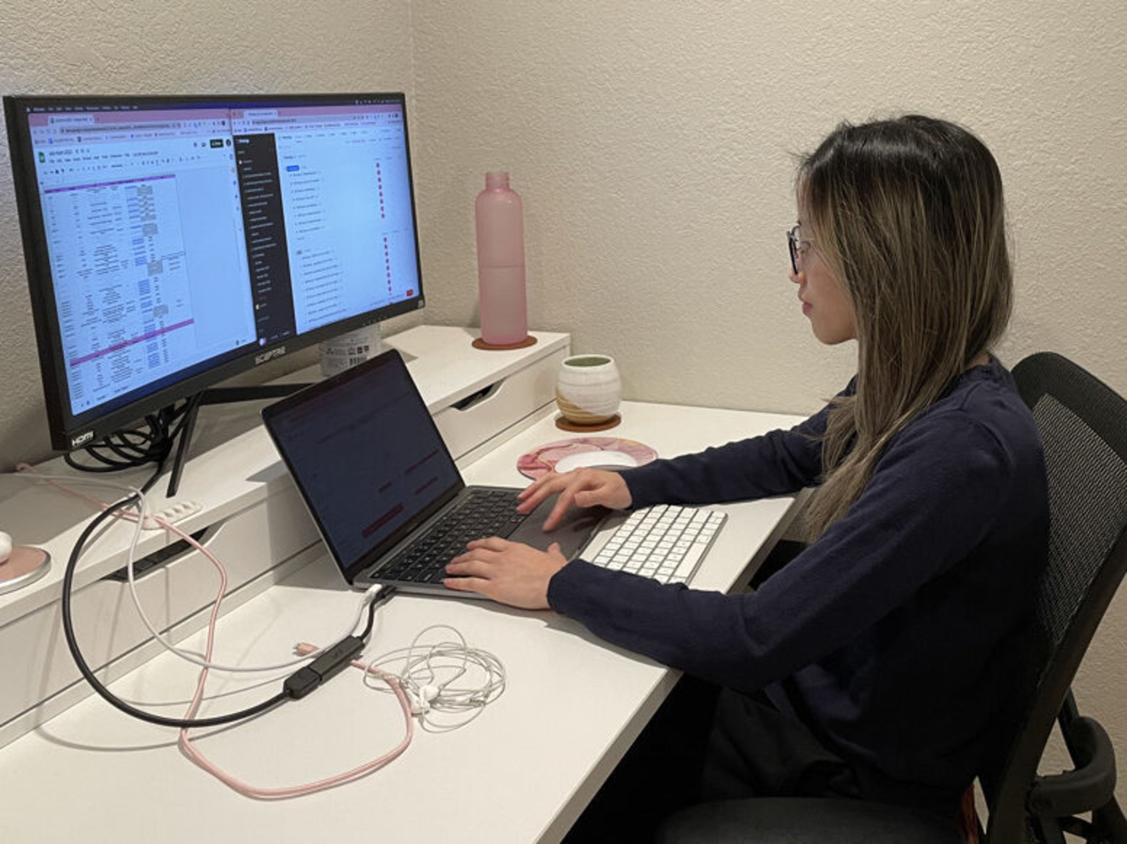A woman with glasses works on a laptop connected to a large monitor, with various cables and a keyboard on a white desk in a minimalist room.