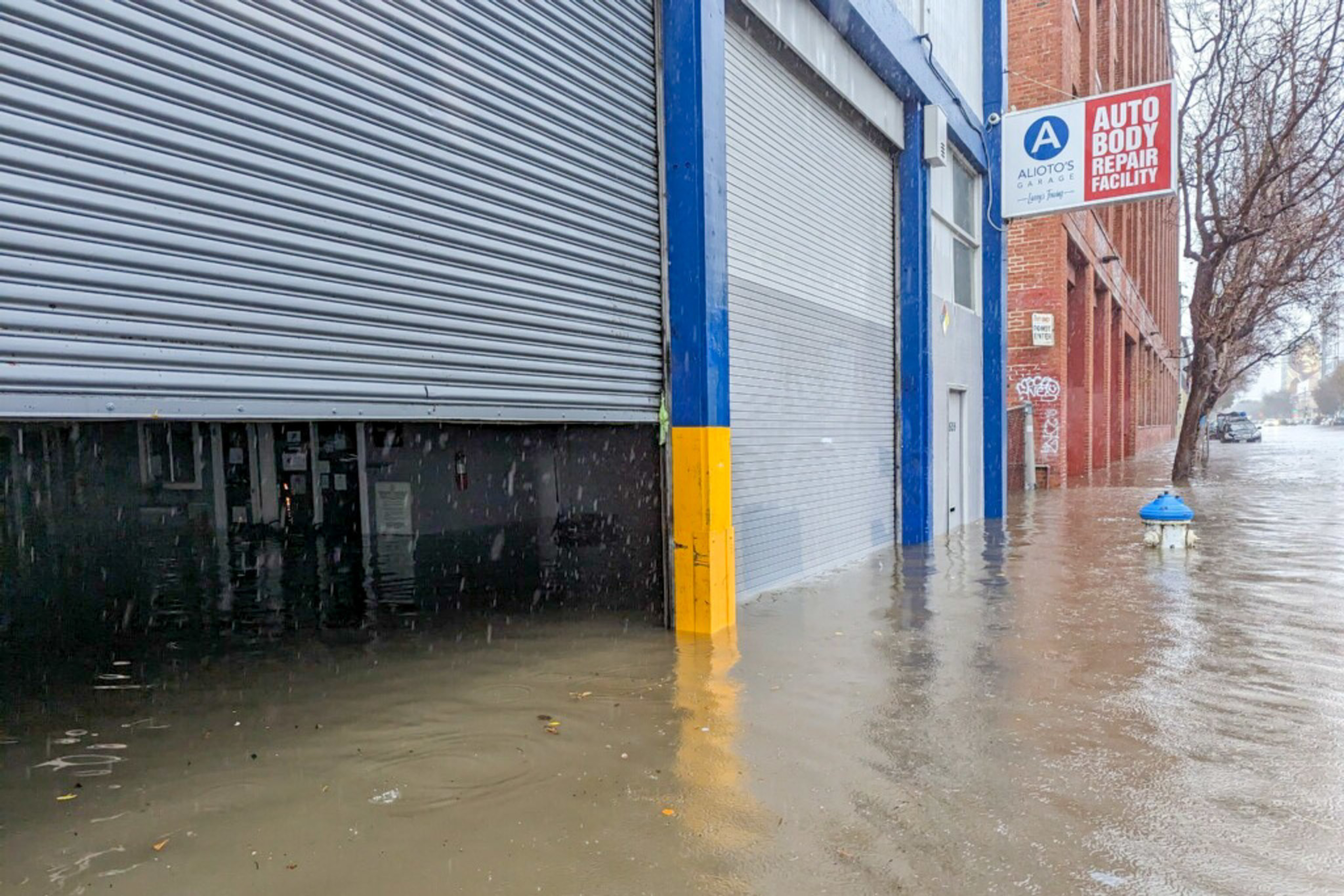 A flooded street with water reaching halfway up garage doors of an auto body repair facility, with a blue fire hydrant partially submerged nearby.