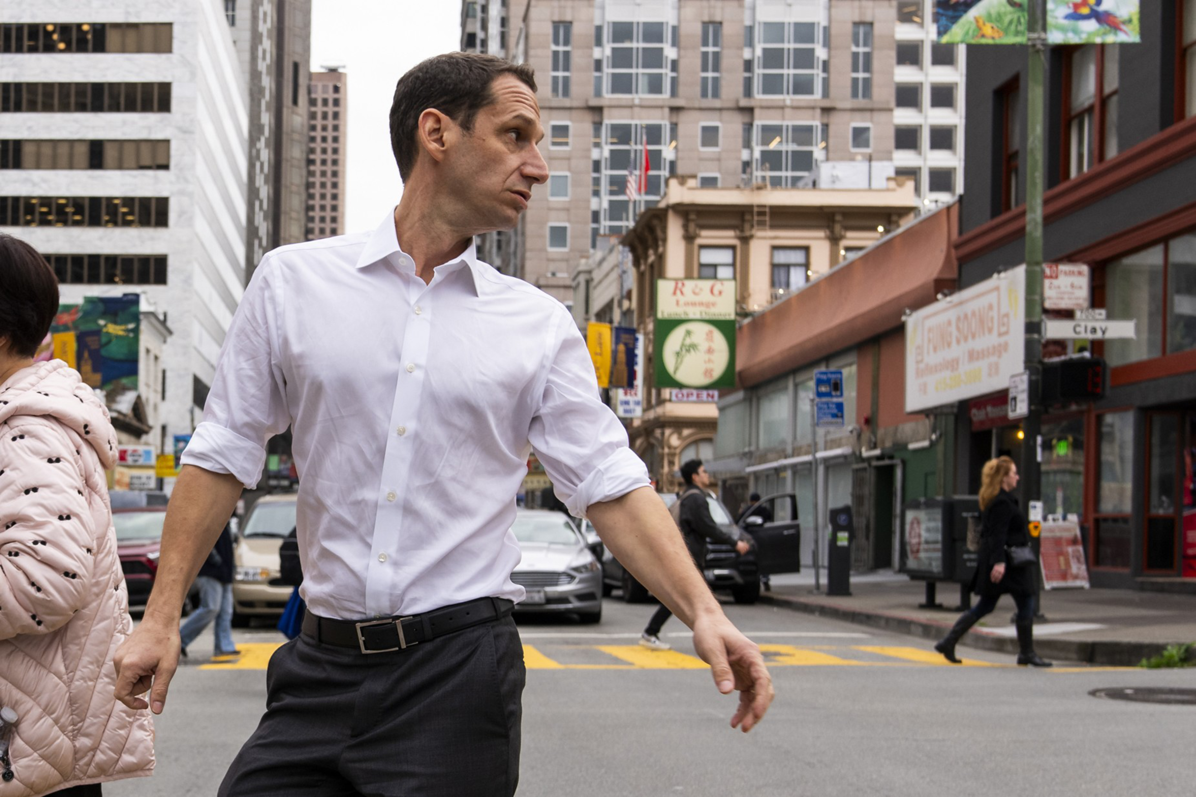 A man in a white shirt crosses a busy city street, turning his head to the side. Buildings, people walking, and cars are visible in the background.