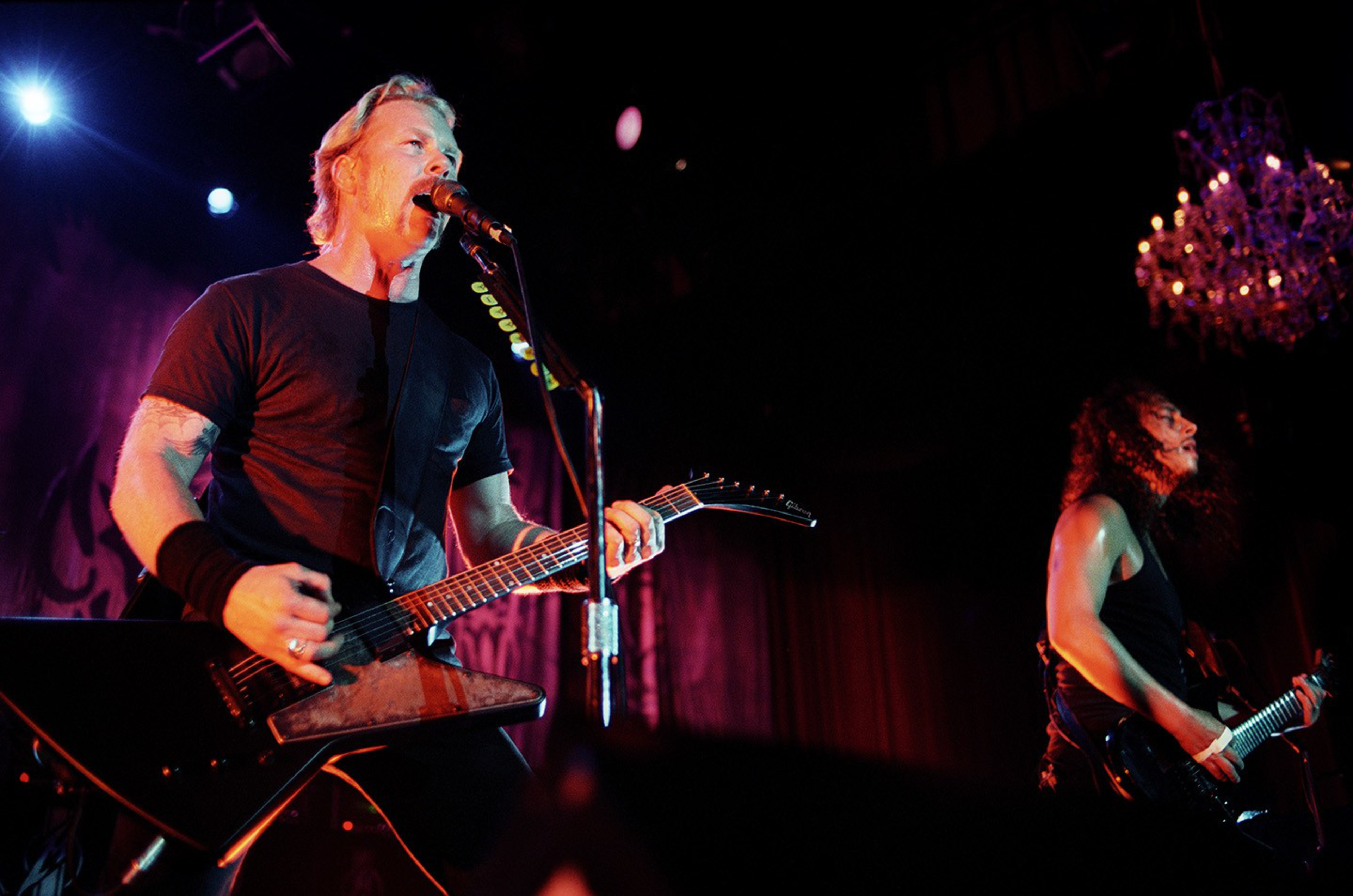 A man with blonde hair sings into a microphone while playing an electric guitar, and another long-haired man plays guitar under warm stage lights.