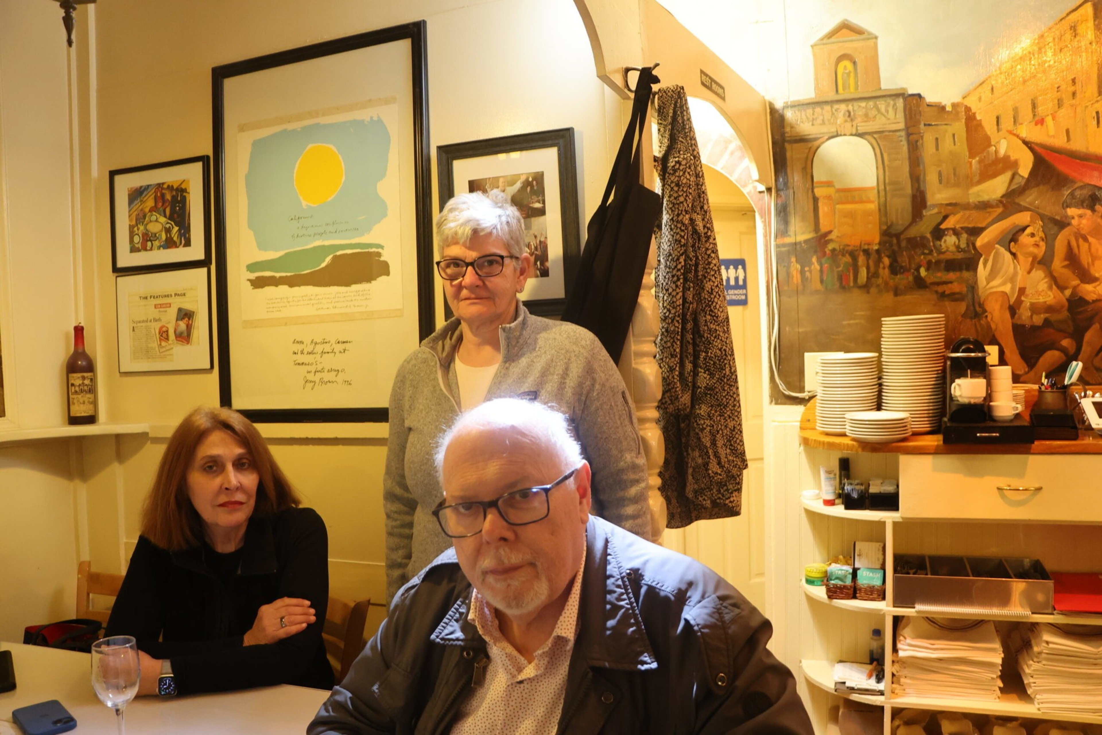 Three adults, two women and one man, are in a warmly lit room with framed art and a mural, with plates and kitchen items on shelves nearby.
