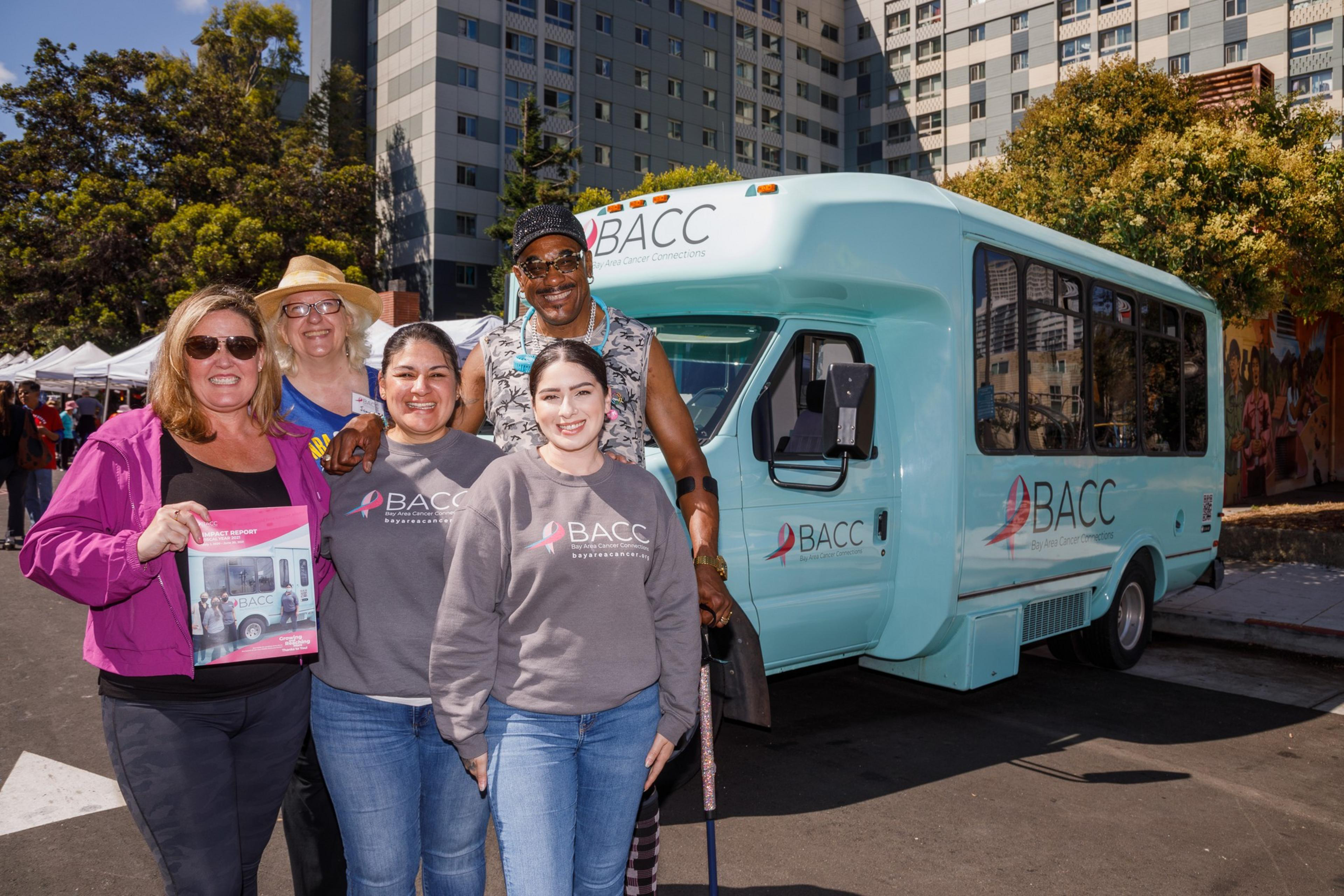 Five people pose by a teal bus with &quot;BACC&quot; logos, at a community event with booths in the background.
