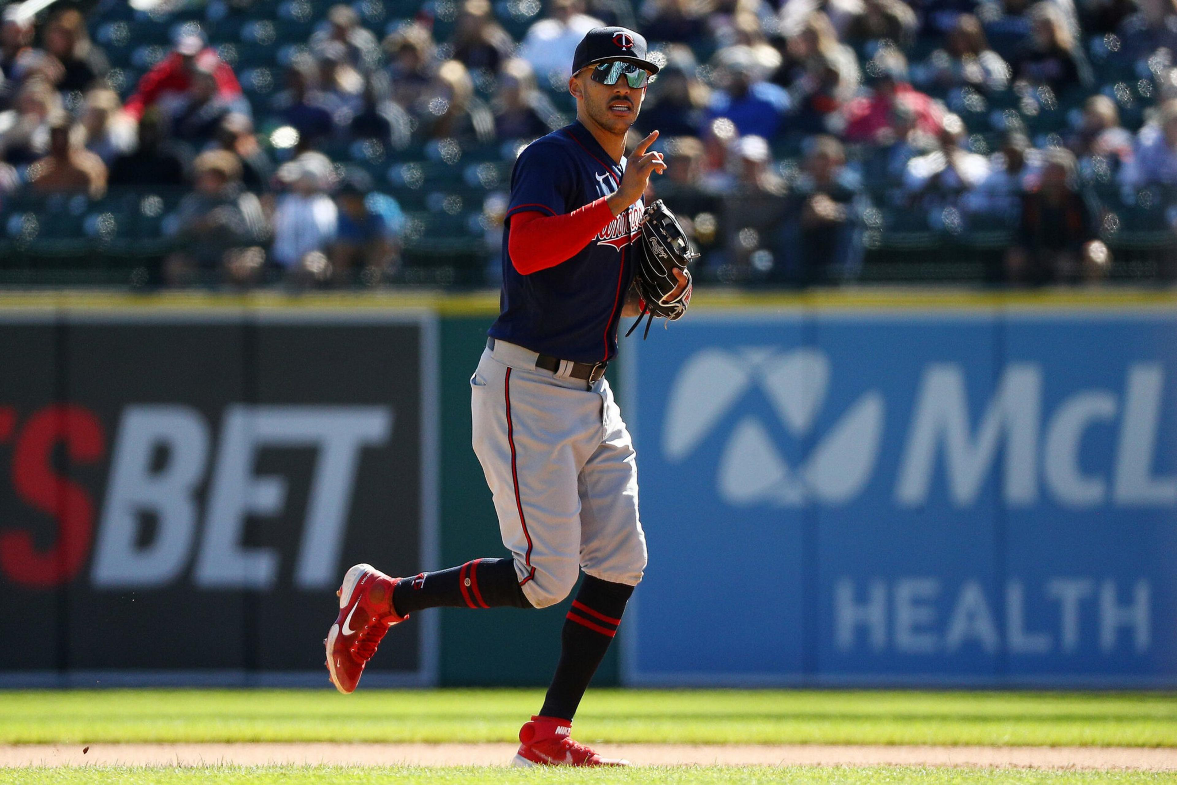 A baseball player in a navy and red uniform signals with one finger while standing on the field, wearing sunglasses and a glove.