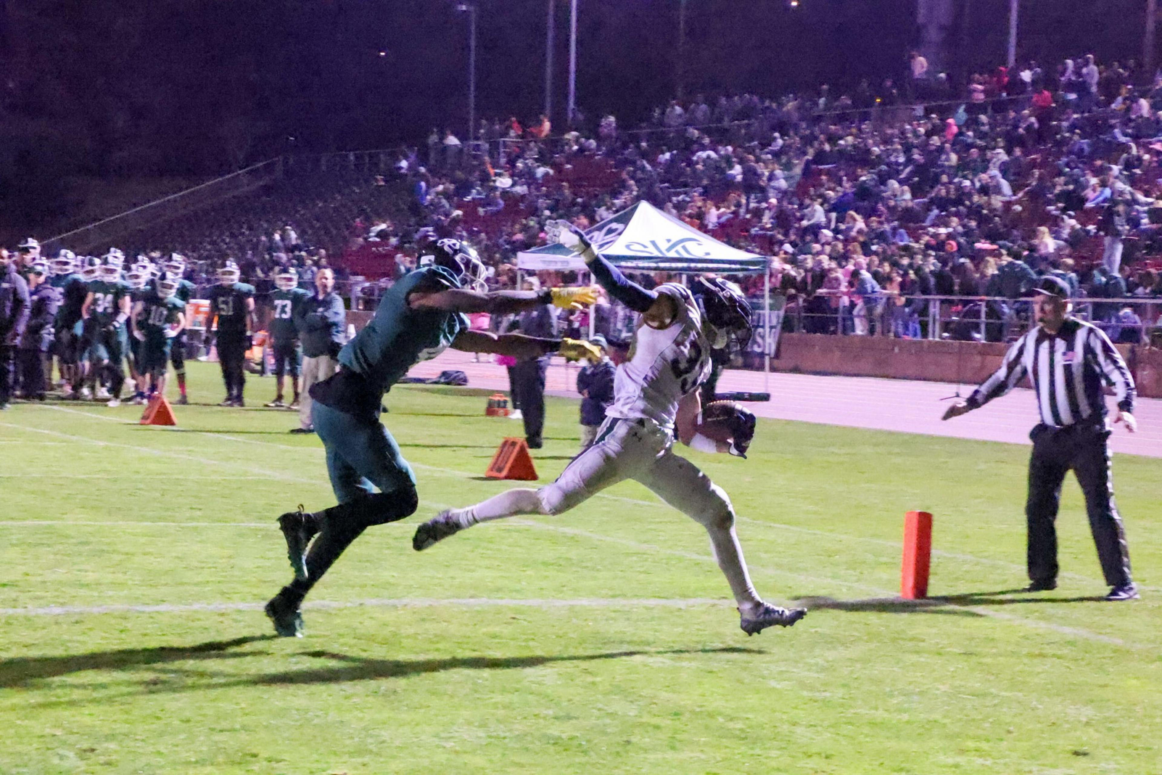 A football player in white stretches toward the end zone, closely pursued by a player in dark green with arms extended, as a referee watches nearby.