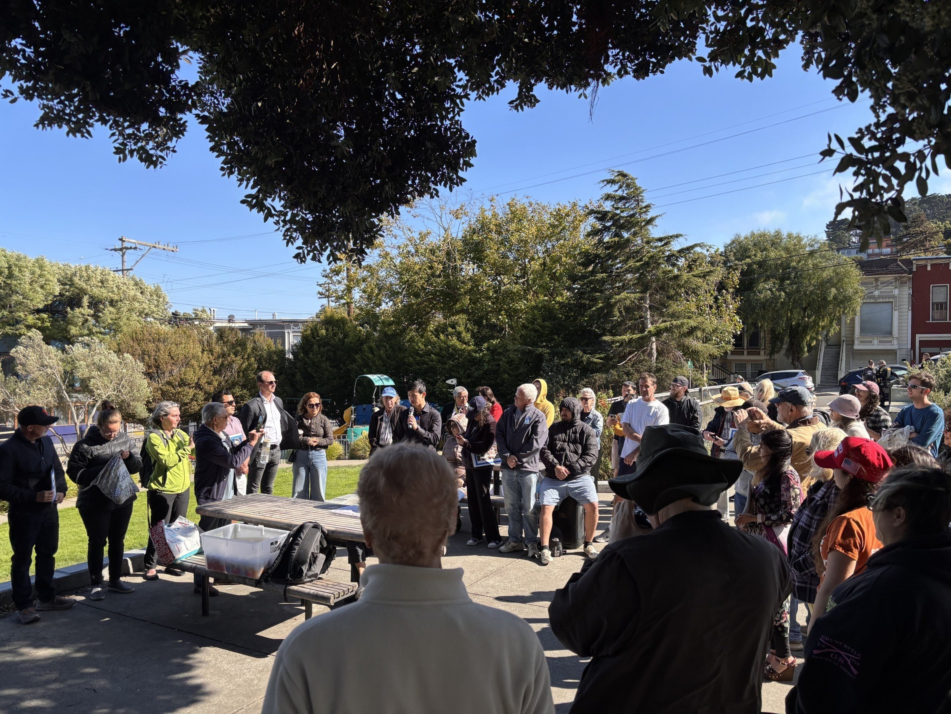 A diverse group of people stands outdoors in a semicircle around a table, listening attentively under clear blue skies and shaded tree branches.