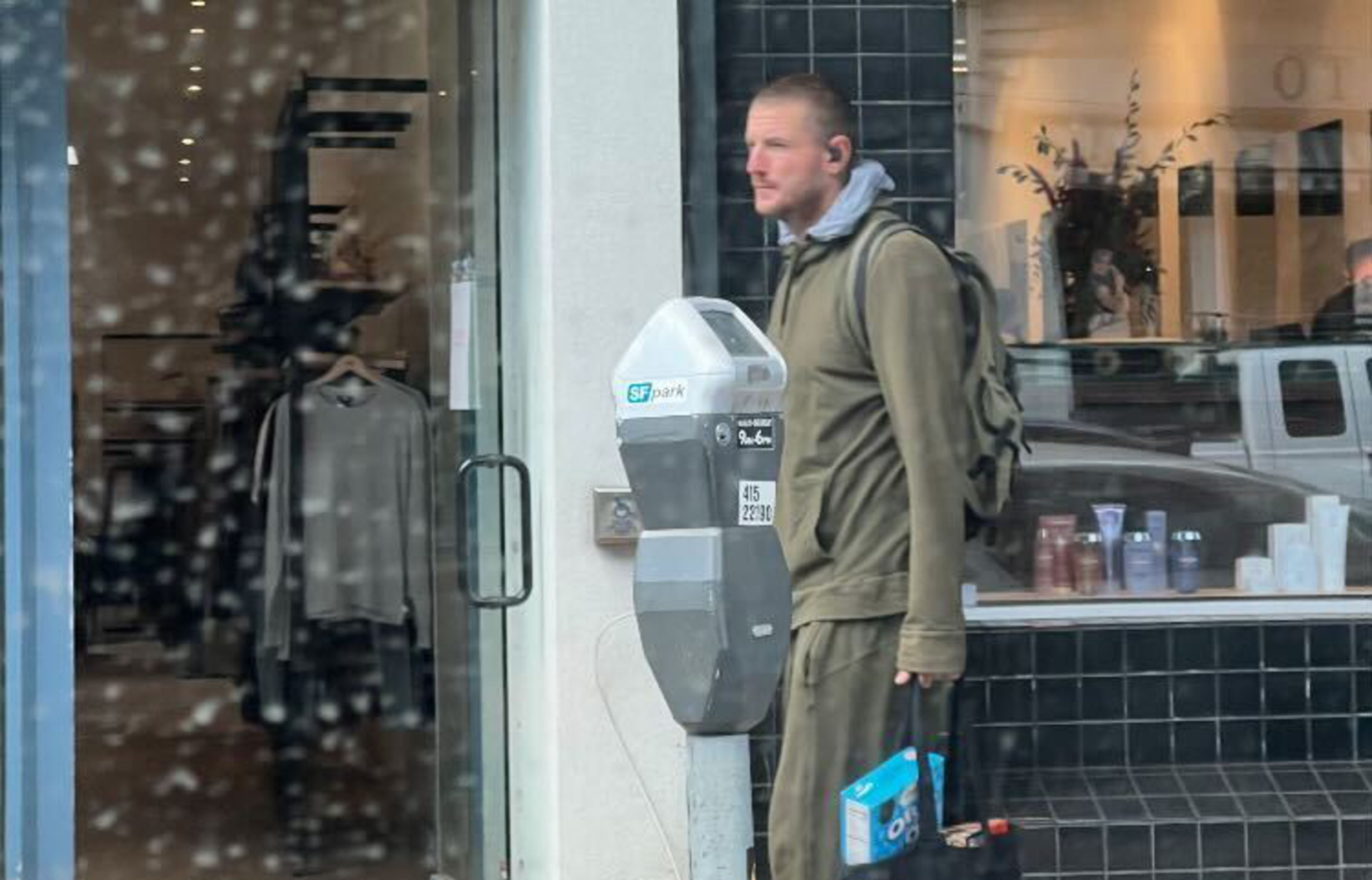 A man in a green tracksuit and backpack stands next to a parking meter outside a store, holding a black bag with a cereal box inside.