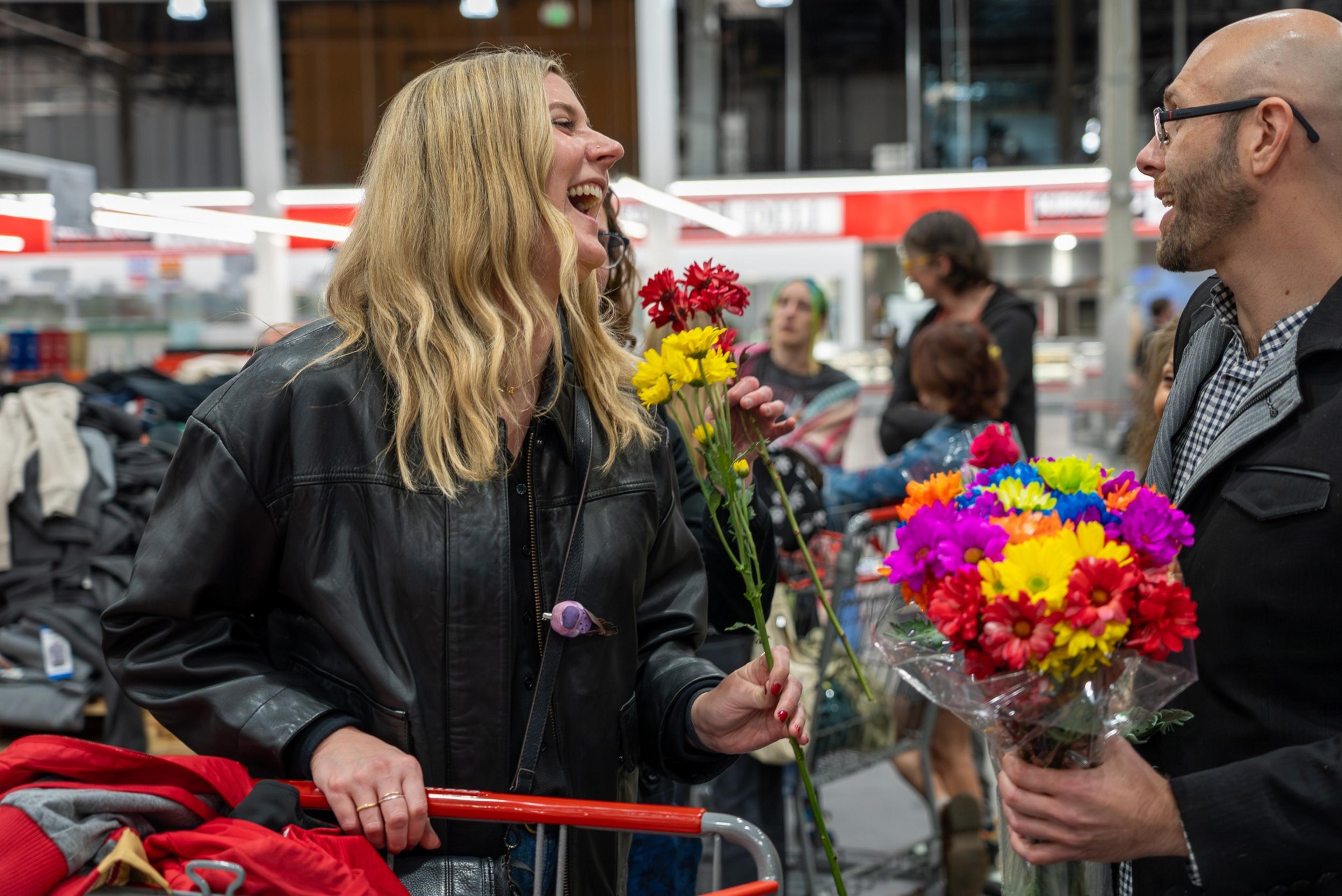 A smiling woman holds red and yellow flowers while talking to a man holding a colorful bouquet inside a busy store.