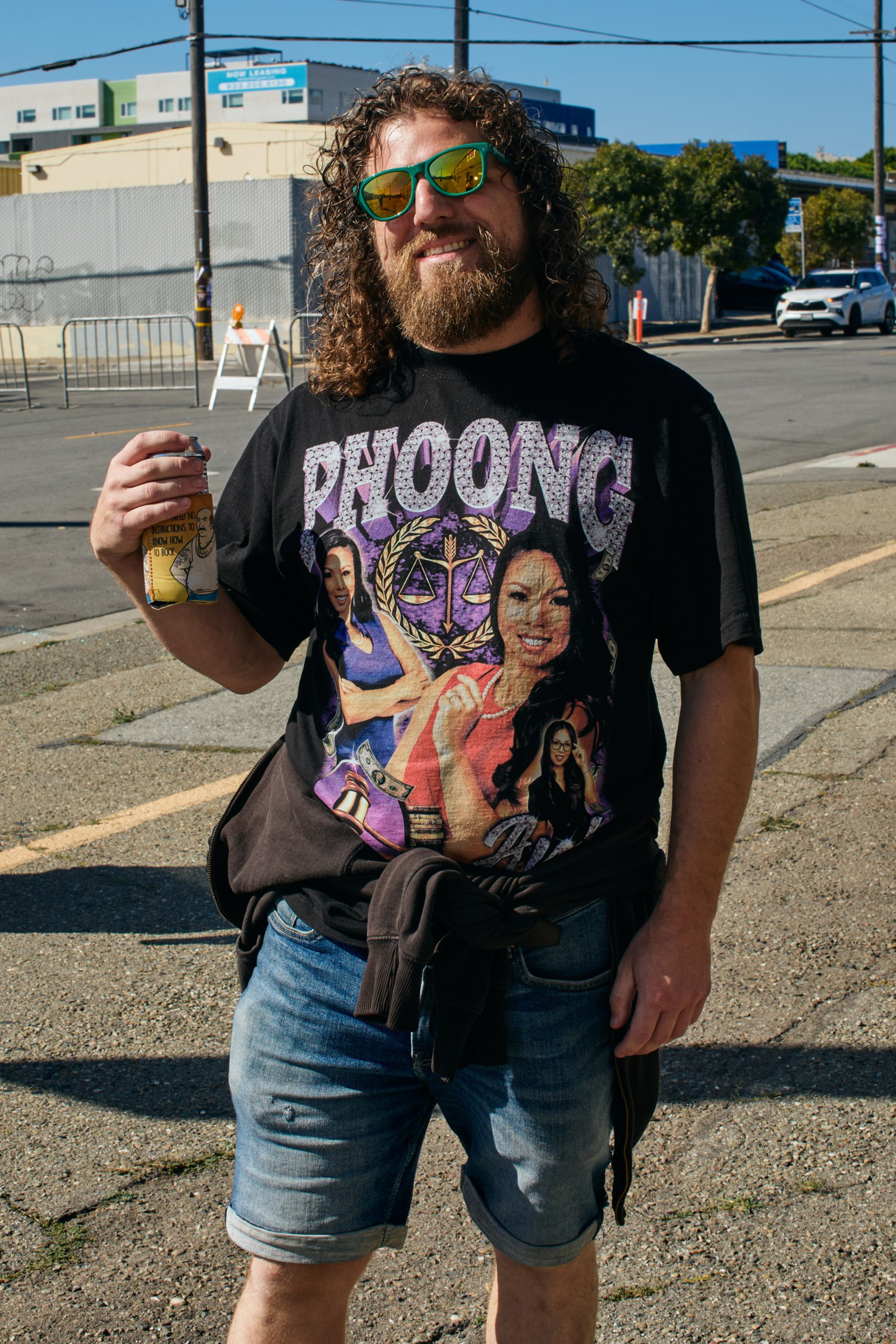 A man with curly hair and a beard wears reflective green sunglasses, a black “PHOONG” graphic tee, denim shorts, and holds a canned drink while smiling.
