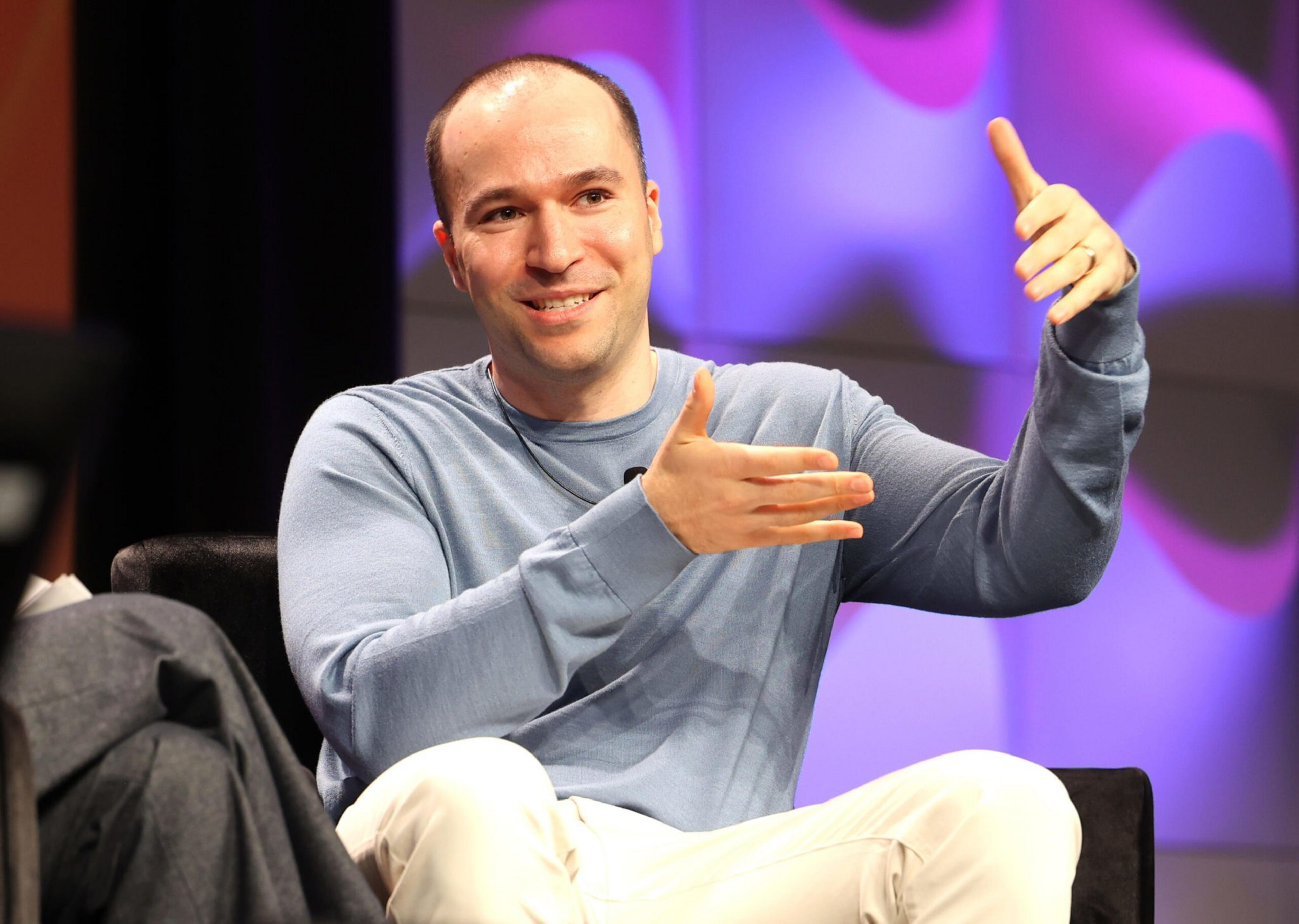 A man in a light blue long-sleeve shirt and white pants sits on a chair, gesturing with both hands while smiling, against a colorful blurred background.