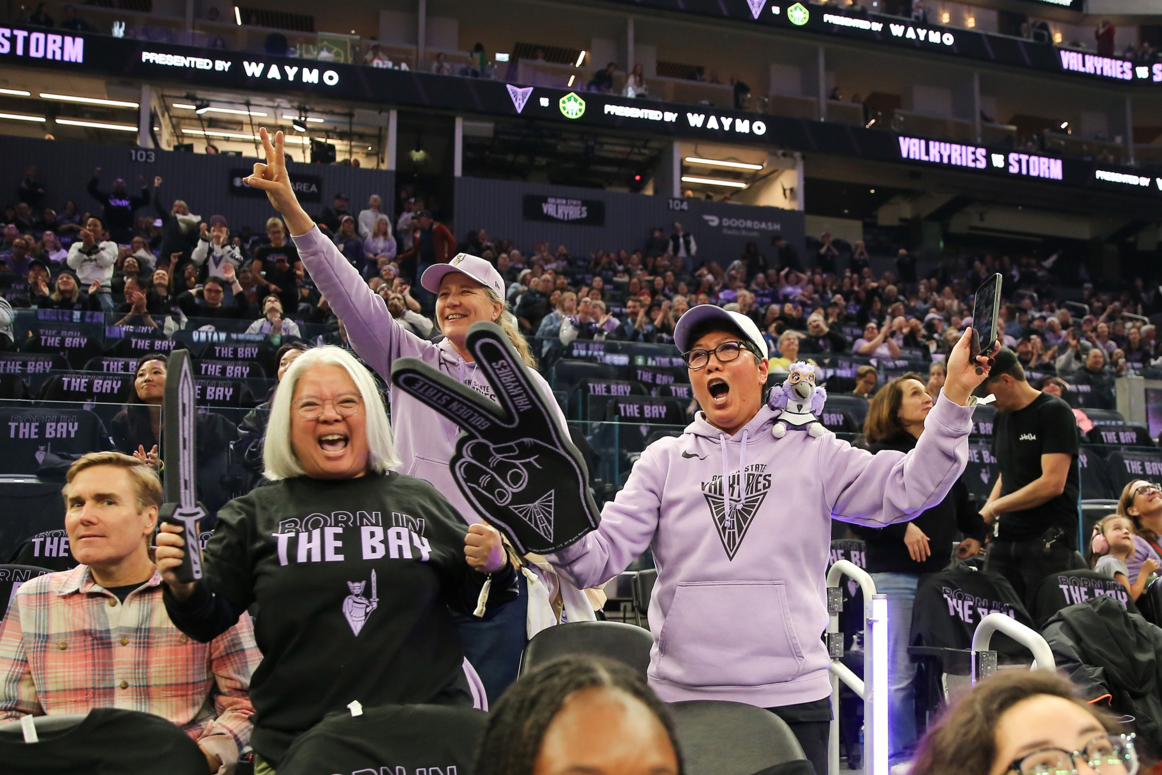 Fans cheer enthusiastically at a sports event, wearing “The Bay” and “Valkyries” gear, holding foam fingers and smiling in a crowded arena.