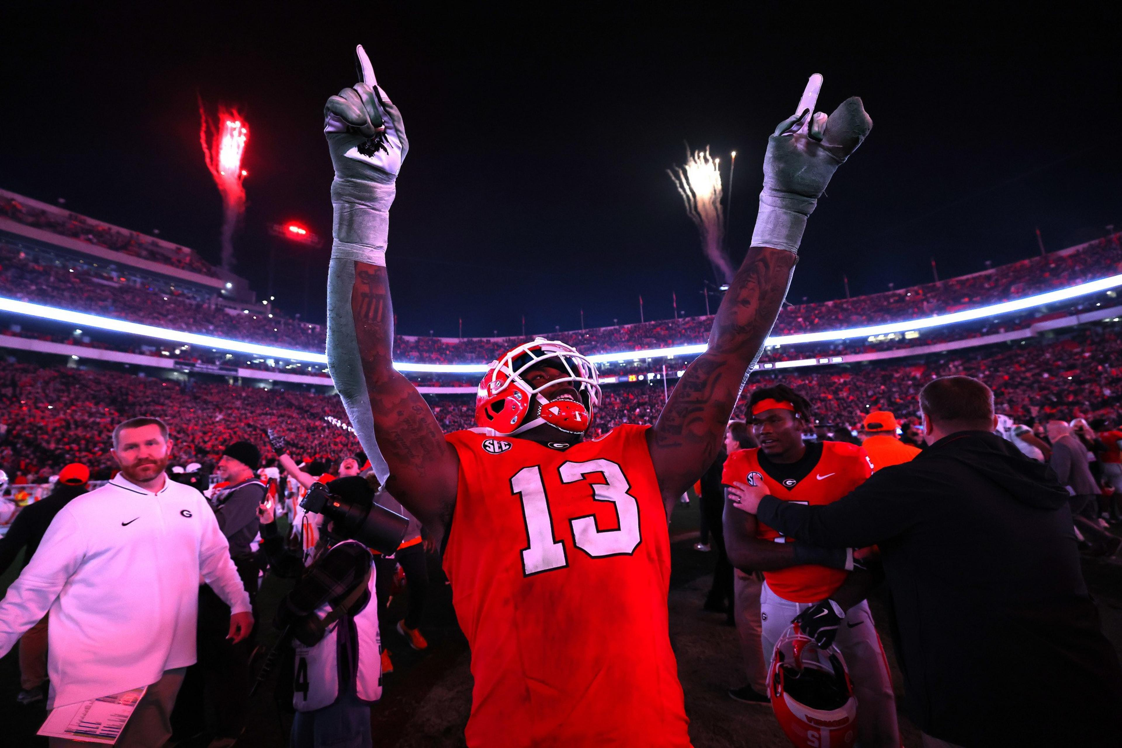 A football player in a red jersey raises his arms in celebration on a field, with fireworks in the night sky and a crowd in the background.