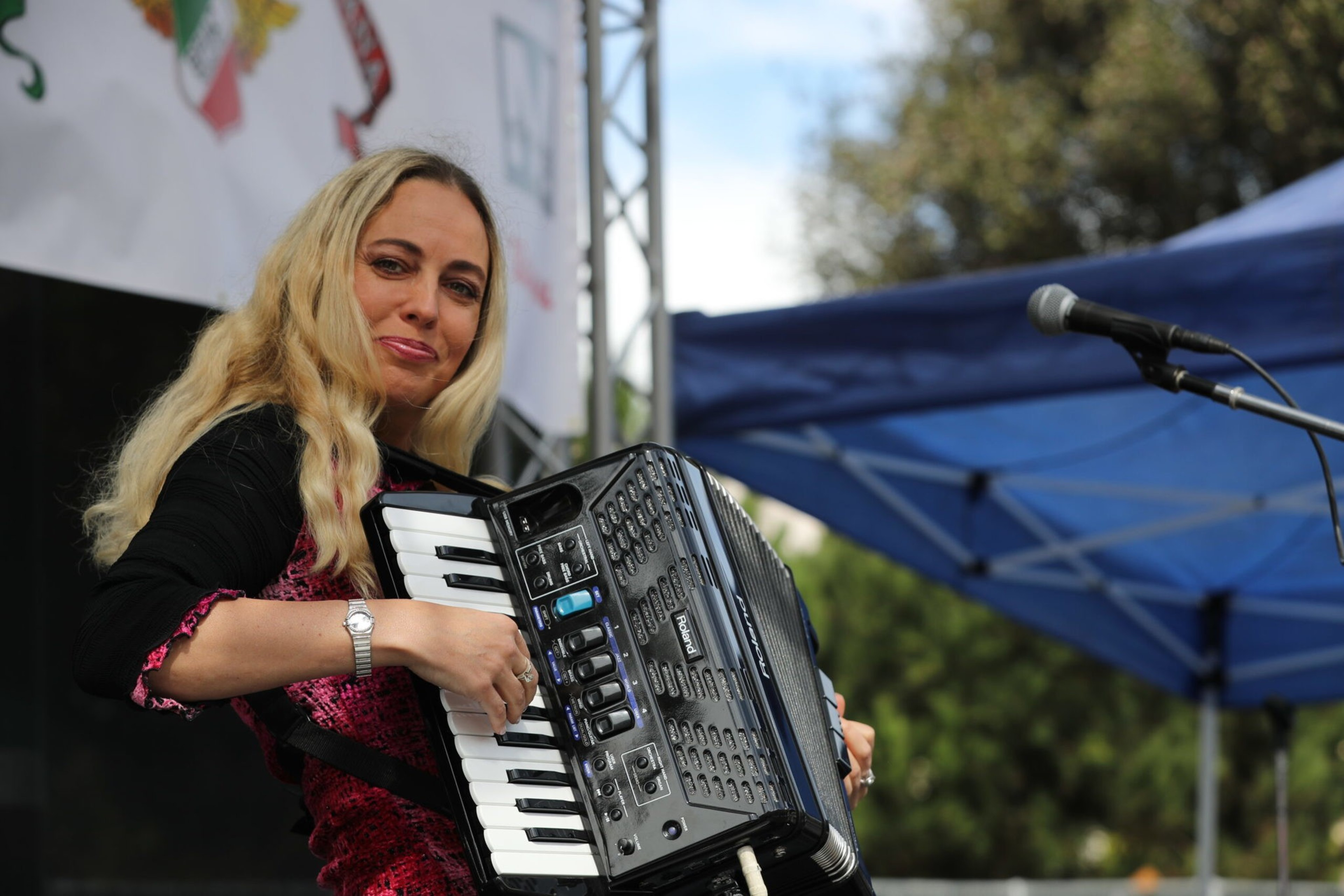A woman with long blonde hair plays a black accordion on an outdoor stage, smiling slightly, with a microphone and blue canopy nearby.