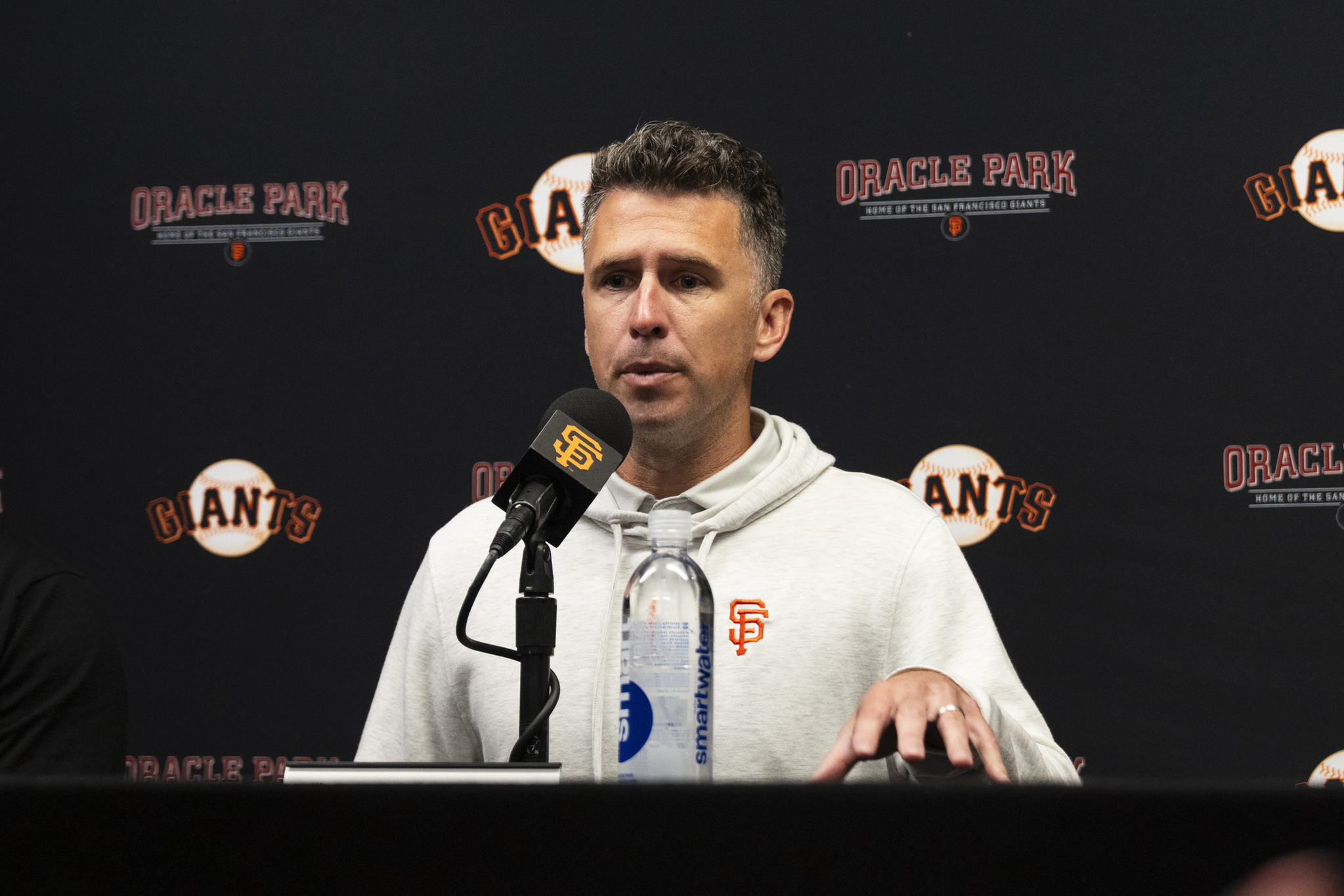 A man in a white hoodie with the San Francisco Giants logo speaks into a microphone at a press conference with "Oracle Park" and "Giants" logos behind him.
