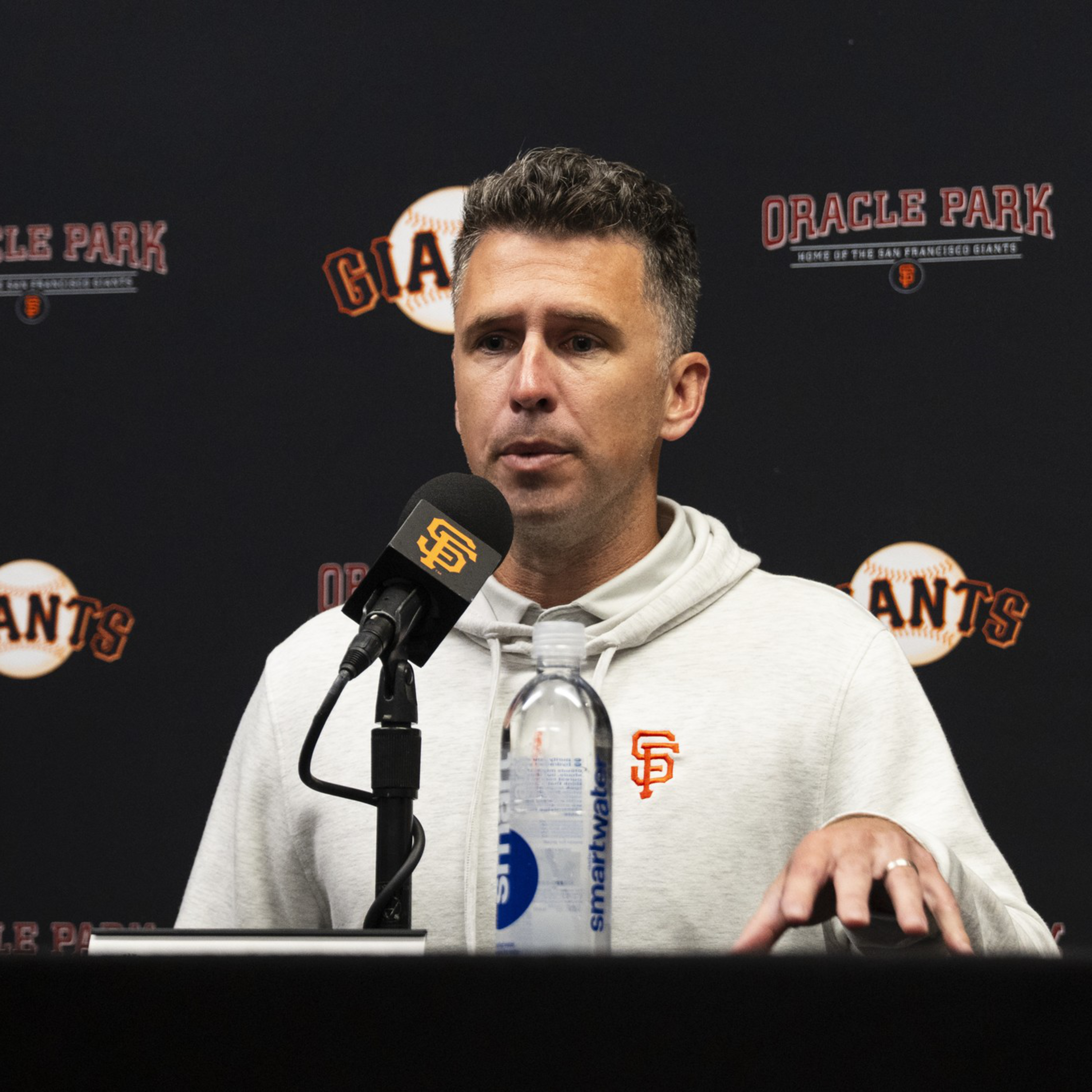 A man in a white hoodie with the San Francisco Giants logo speaks into a microphone at a press conference with "Oracle Park" and "Giants" logos behind him.