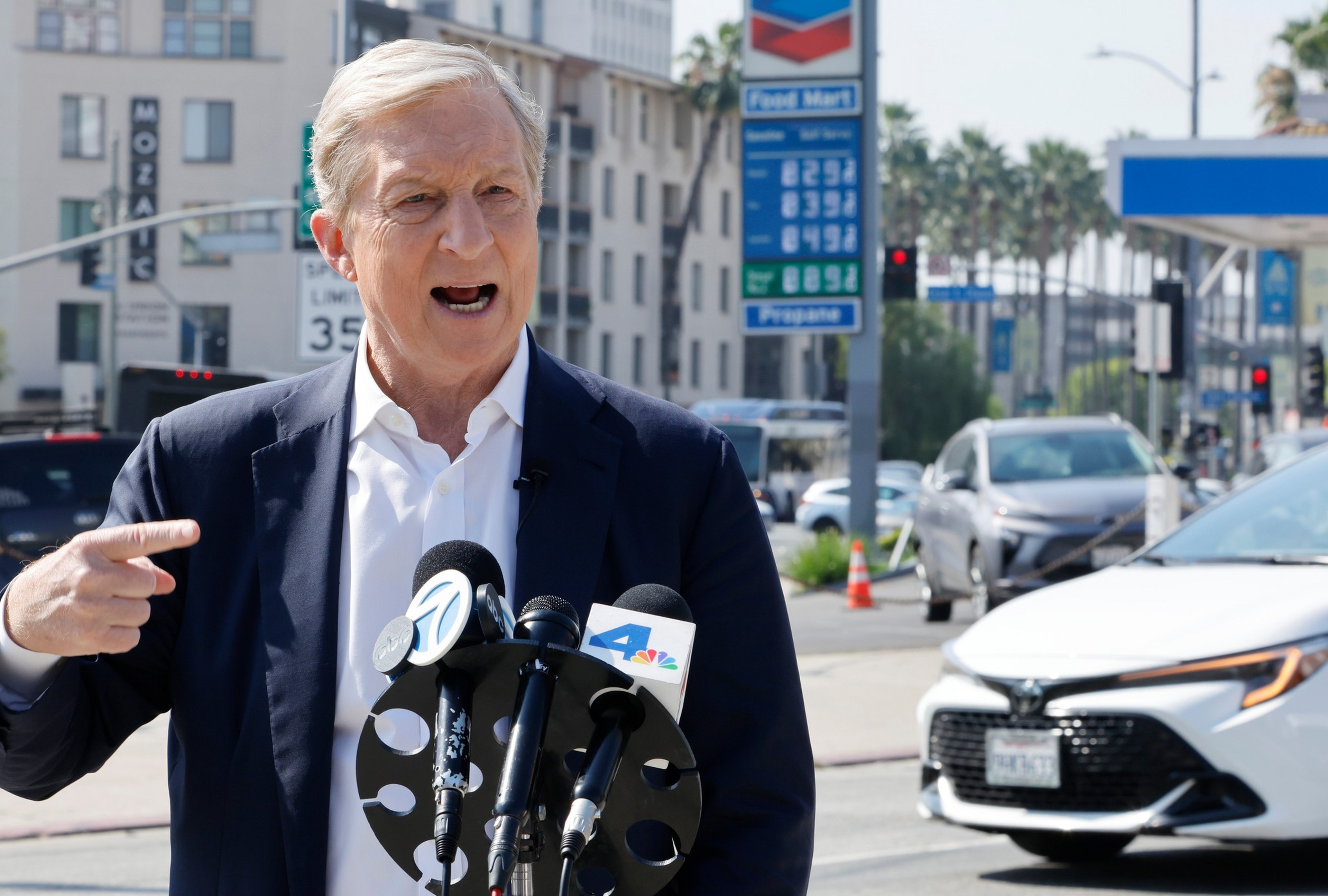 A man in a suit speaks passionately at a press conference on a city street with several microphones in front of him and cars behind.