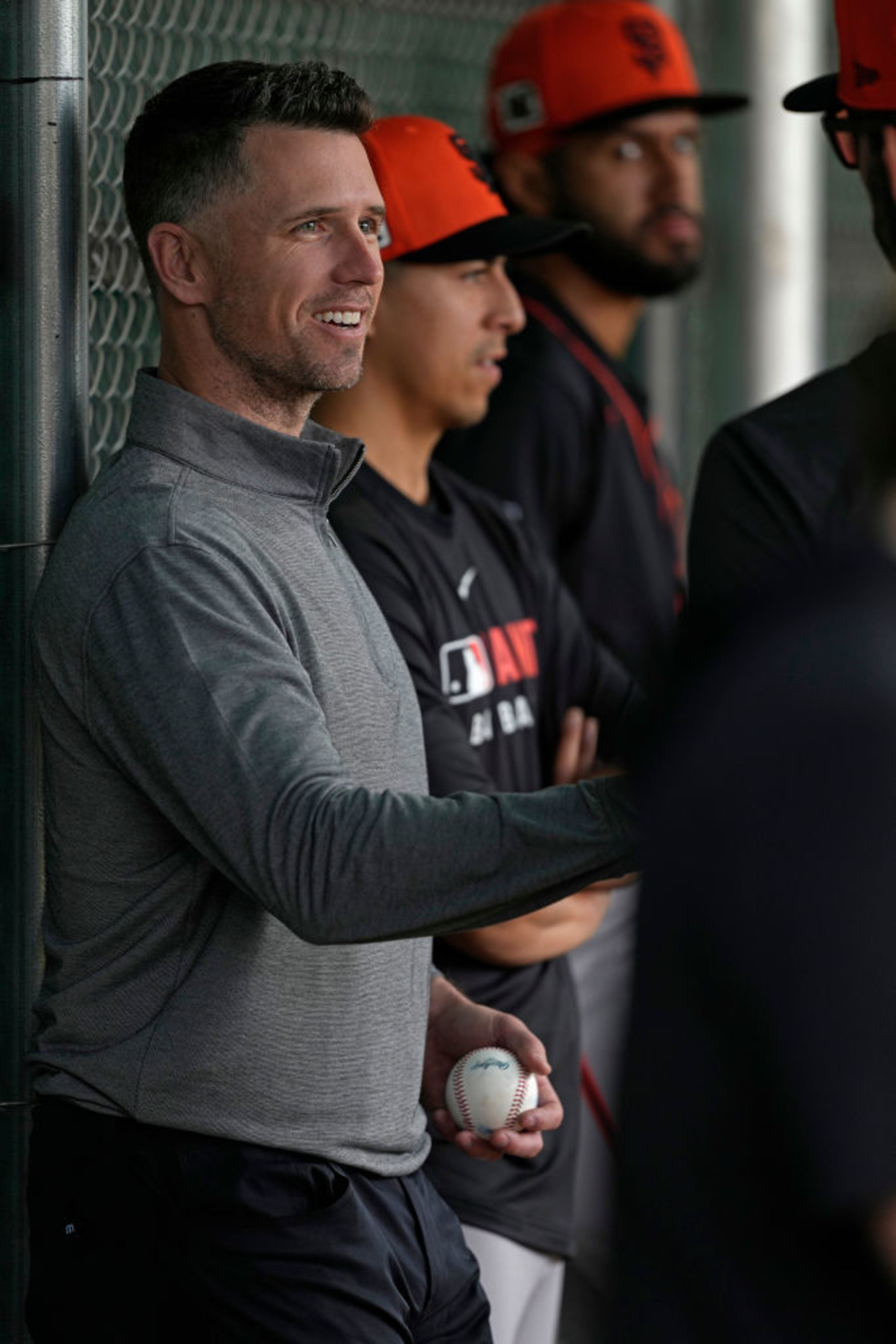 A man in a gray sweater smiles while holding a baseball. He stands beside others wearing black shirts and orange caps, likely at a sports event.