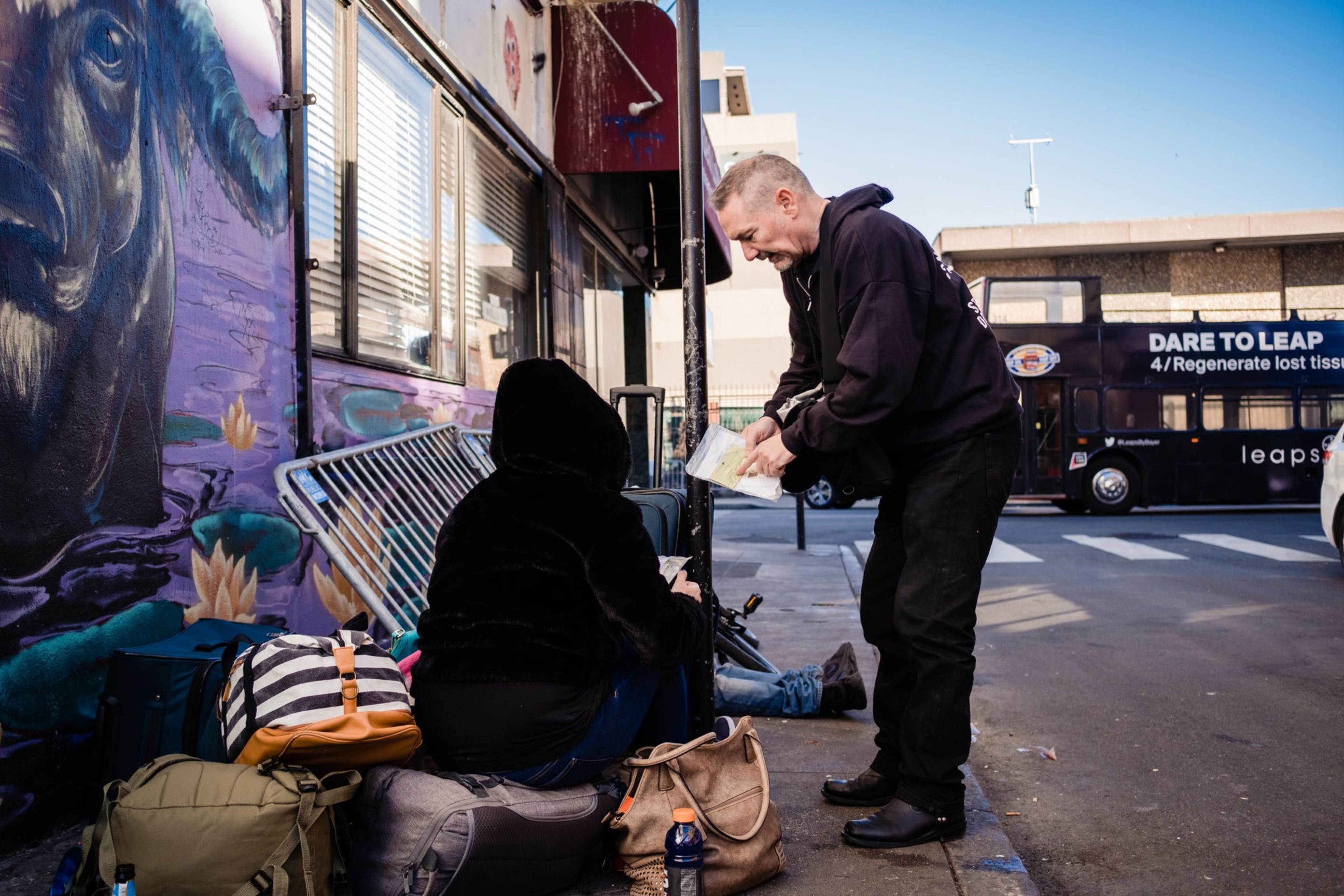 Man holding paper leans over to hooded individual sitting on the sidewalk