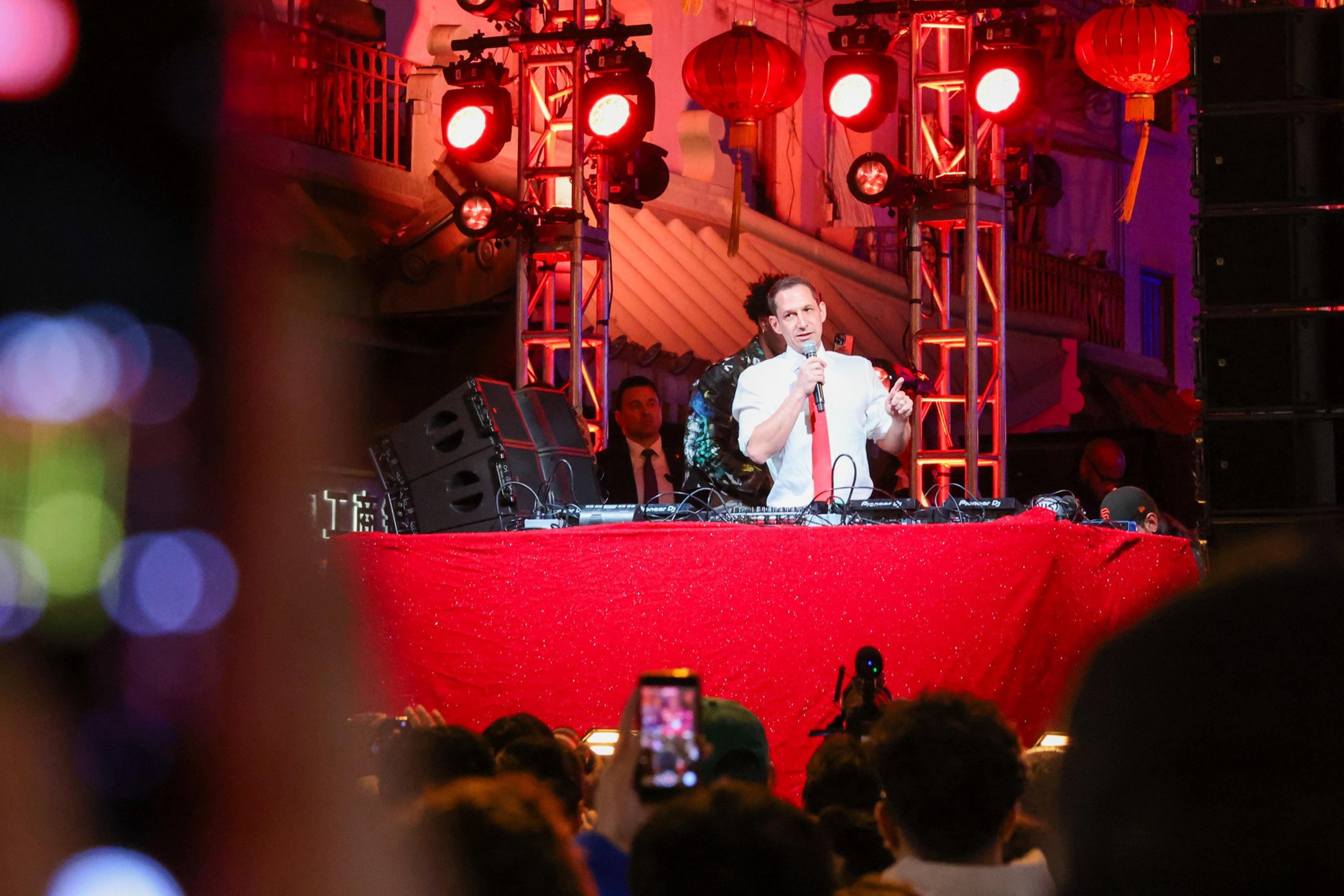 A man in a white shirt and red tie speaks into a microphone behind a red-lit DJ booth as a crowd watches and records with phones.