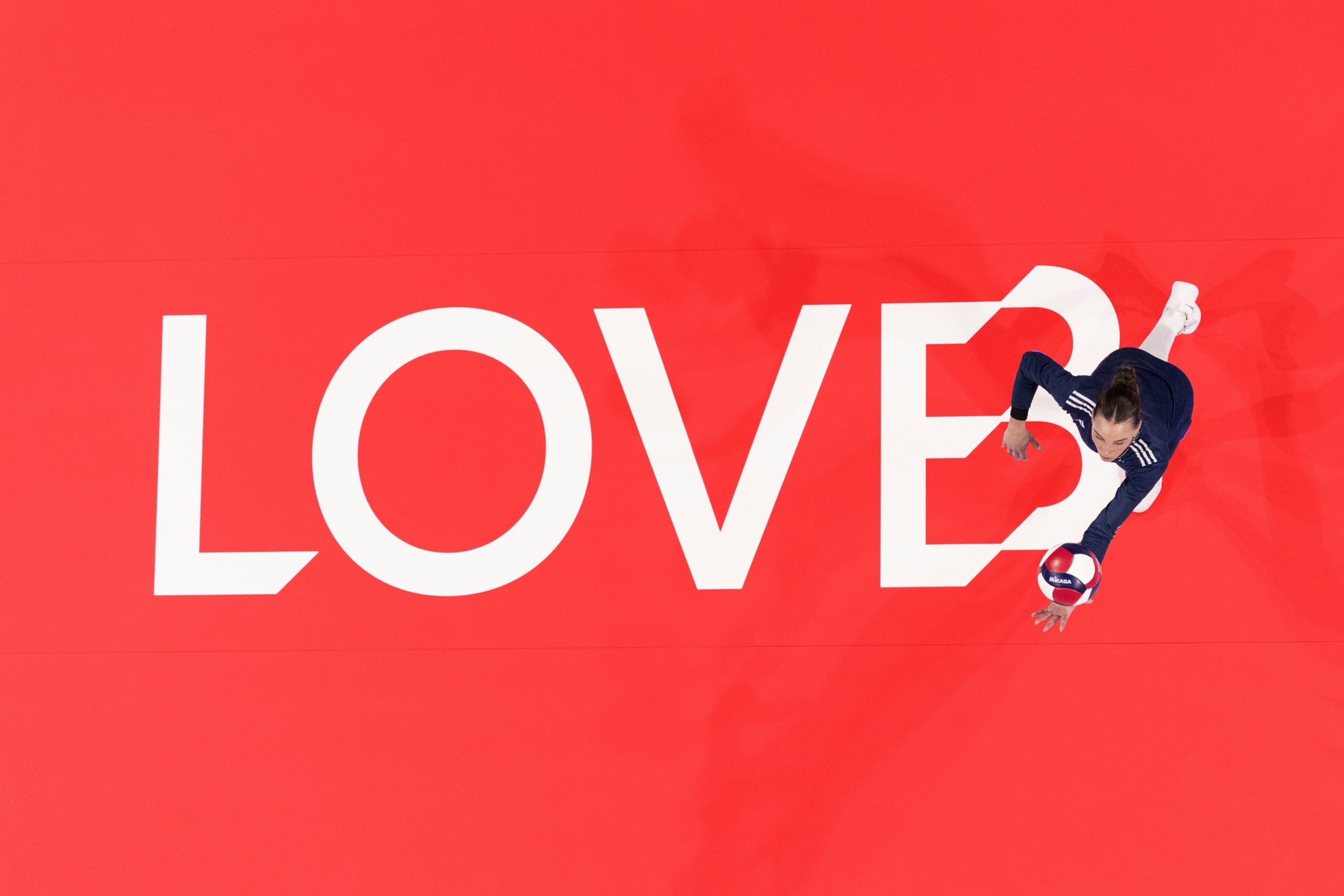 A volleyball player in navy sportswear leaps to hit a ball over a red court with the word “LOVE?” painted in large white letters.