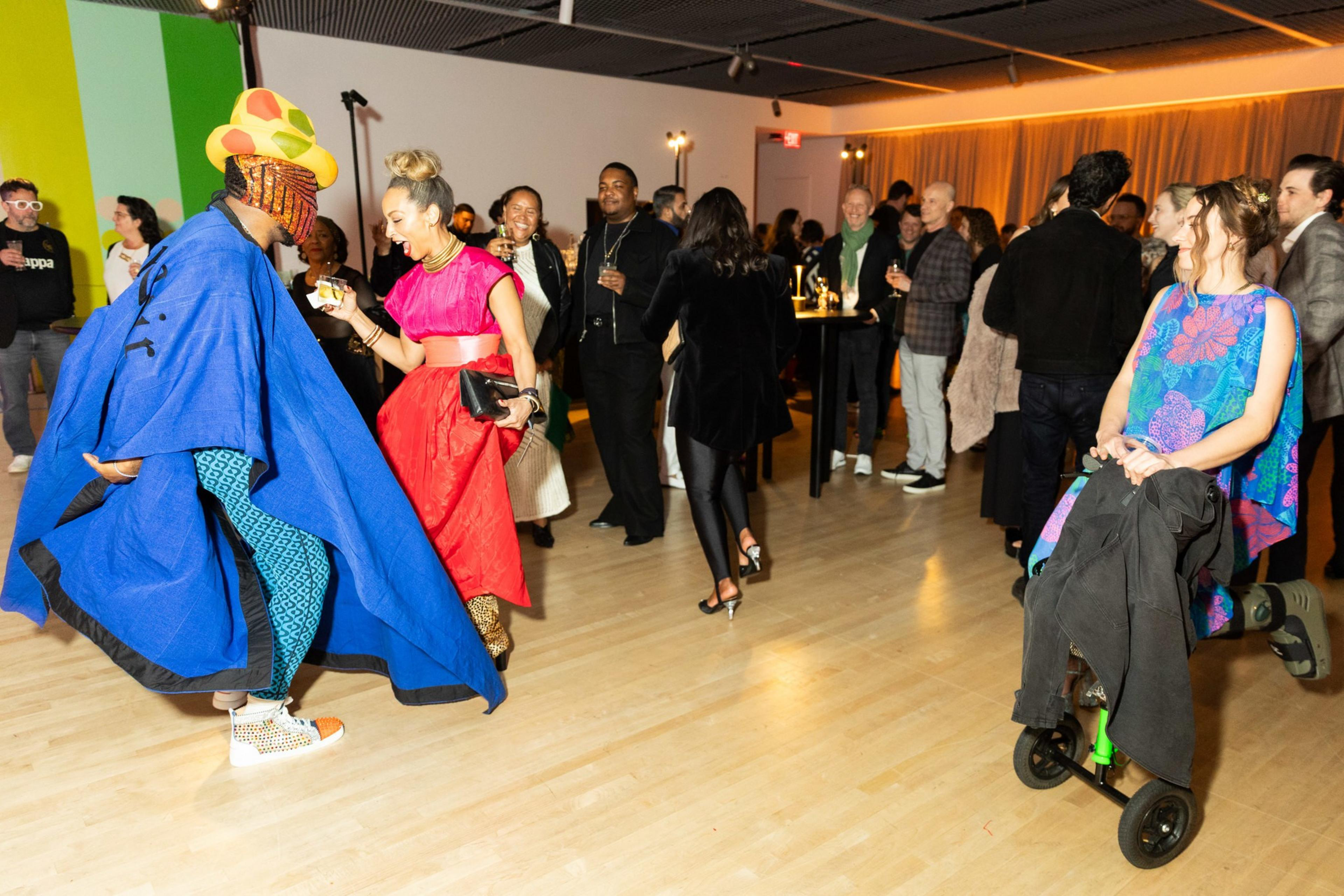 A lively party shows people mingling, dressed in colorful outfits. One person wears a blue cape and patterned clothes, while others smile and engage warmly.