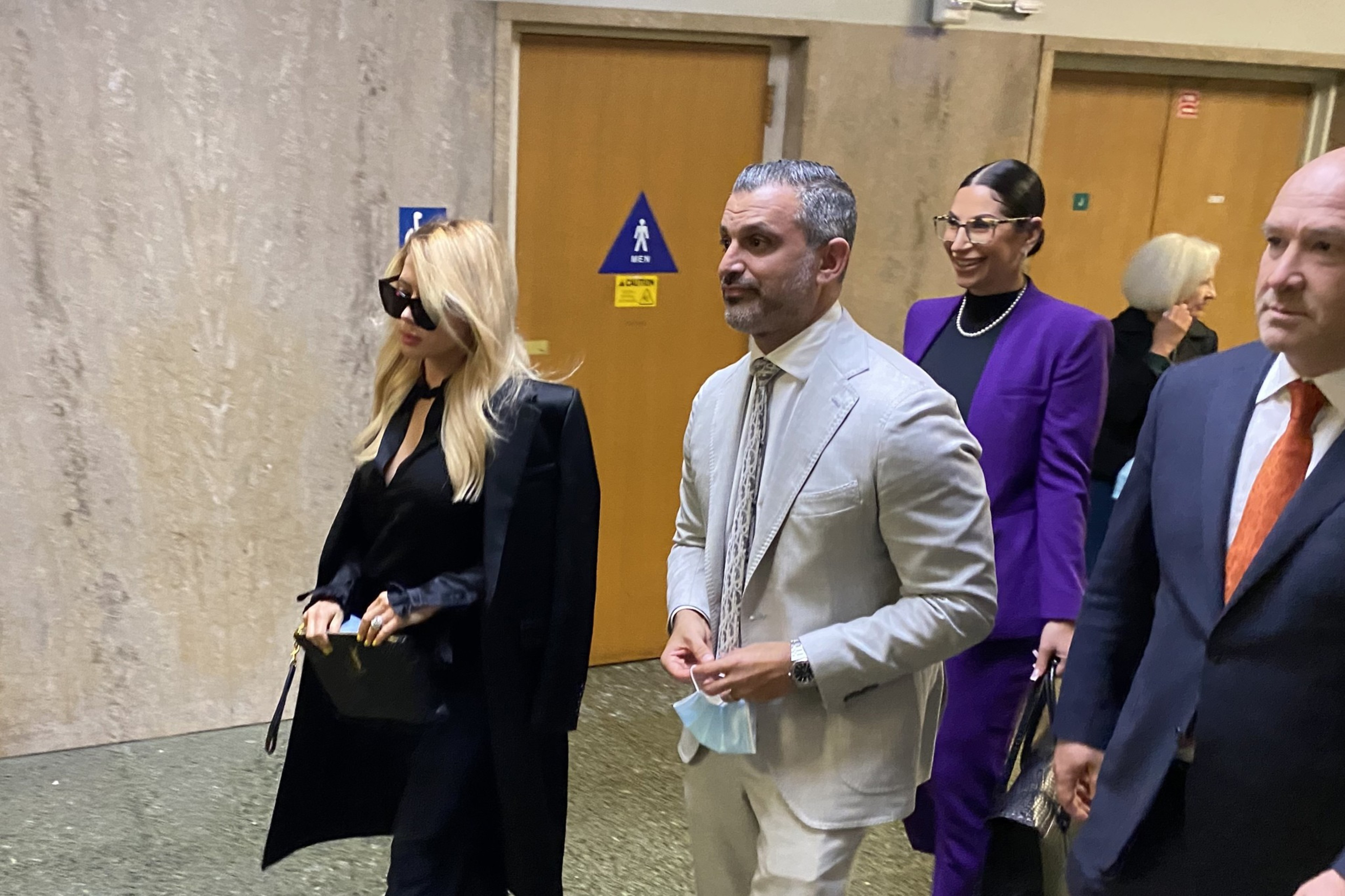 Four well-dressed people walk down a hallway, two women wearing sunglasses and a purple suit, and two men in light gray and dark suits.