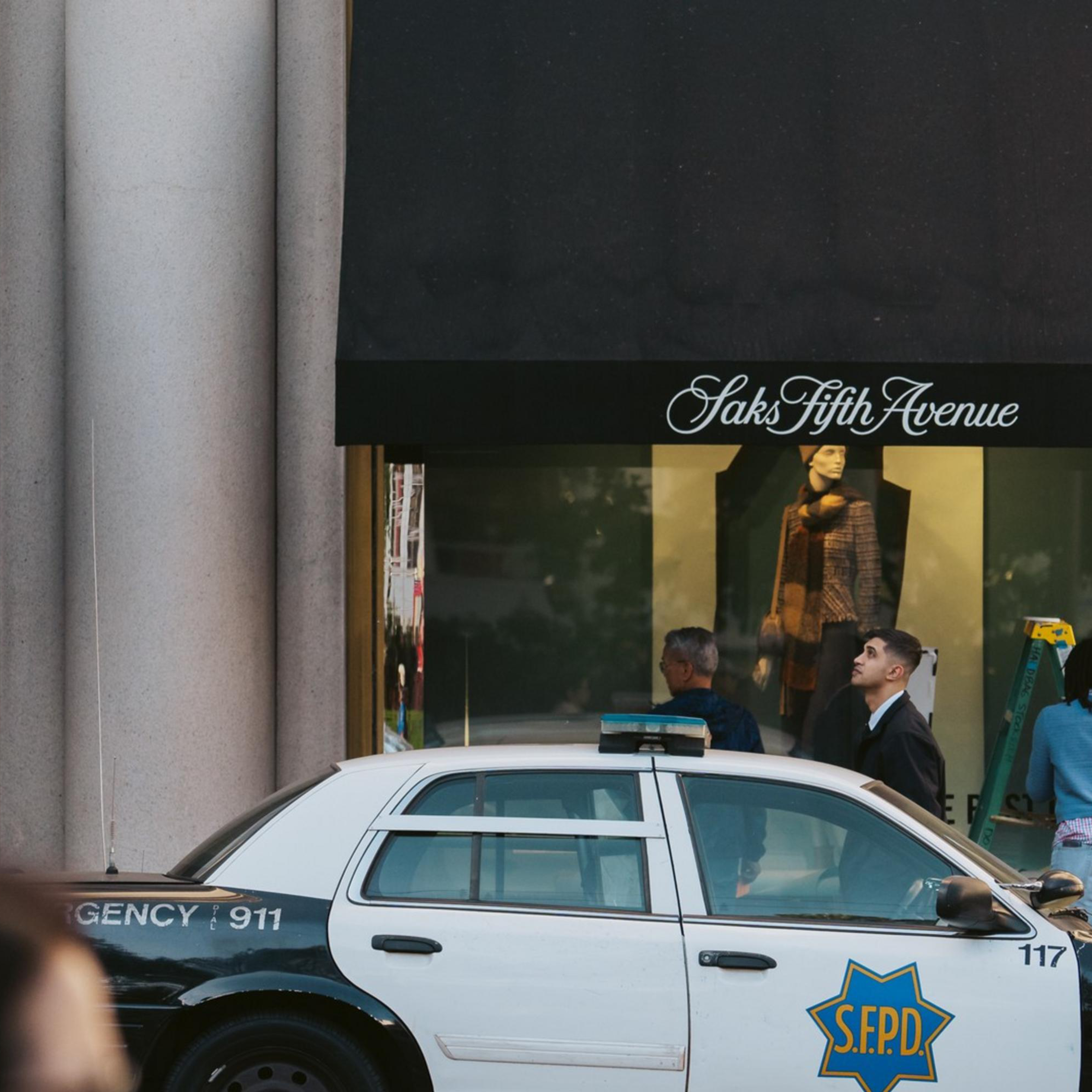 A police car is parked outside a Saks Fifth Avenue store with people visible in front of the windows and a blurred passerby in the foreground.