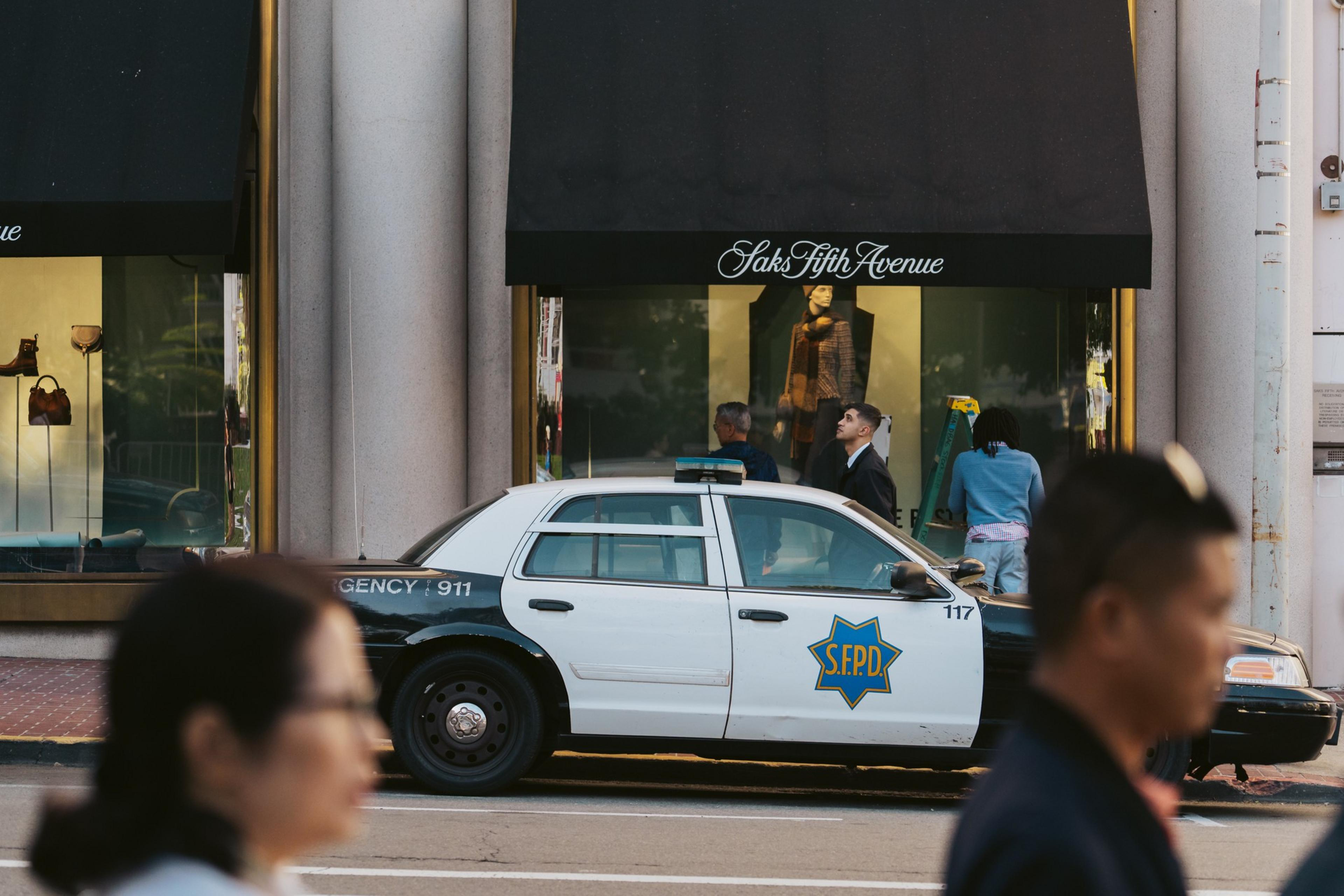 A police car is parked outside a Saks Fifth Avenue store with people visible in front of the windows and a blurred passerby in the foreground.