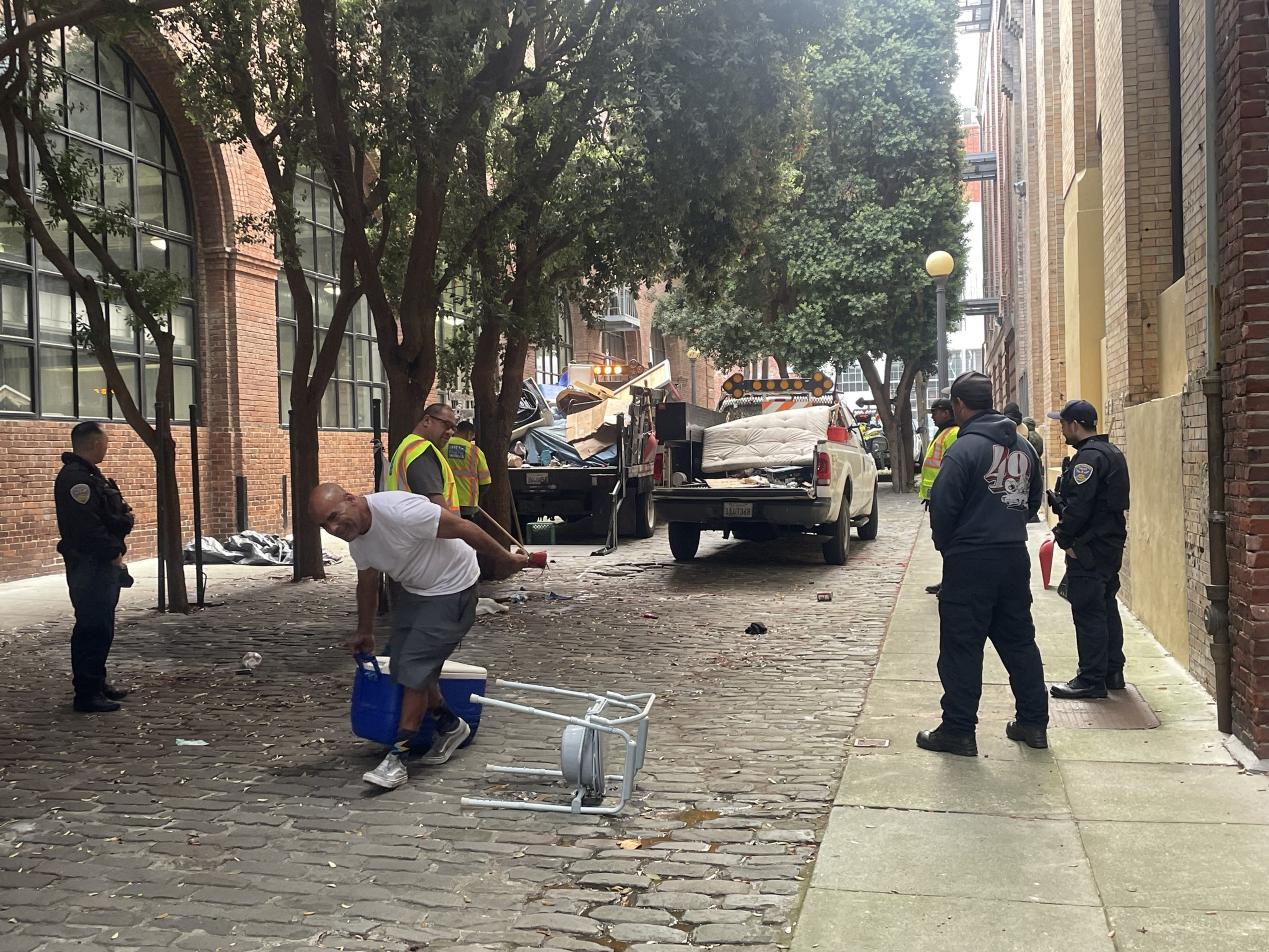 A man drags a cooler as city workers look on. A trash truck filled with the belongings of unhoused people is in the background.