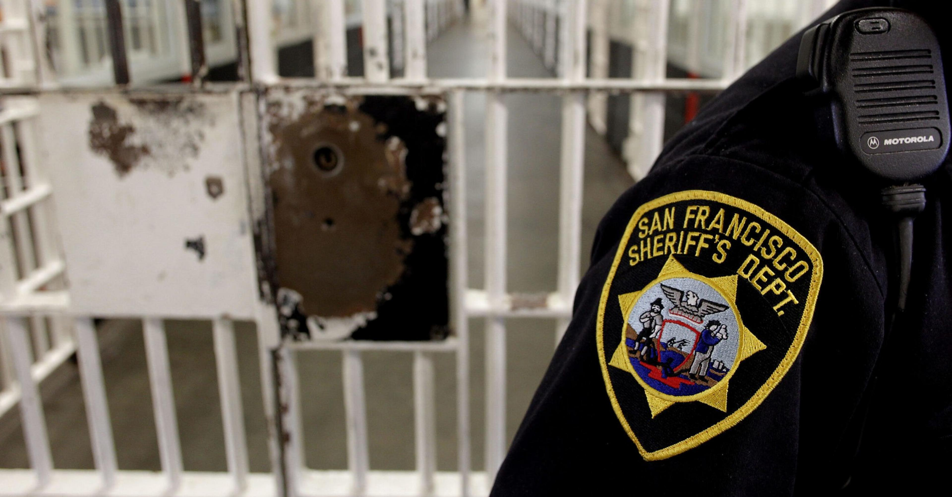 The image shows a San Francisco Sheriff's Dept. badge on an officer's uniform in the foreground, with a worn, metal prison cell door and corridor in the background.