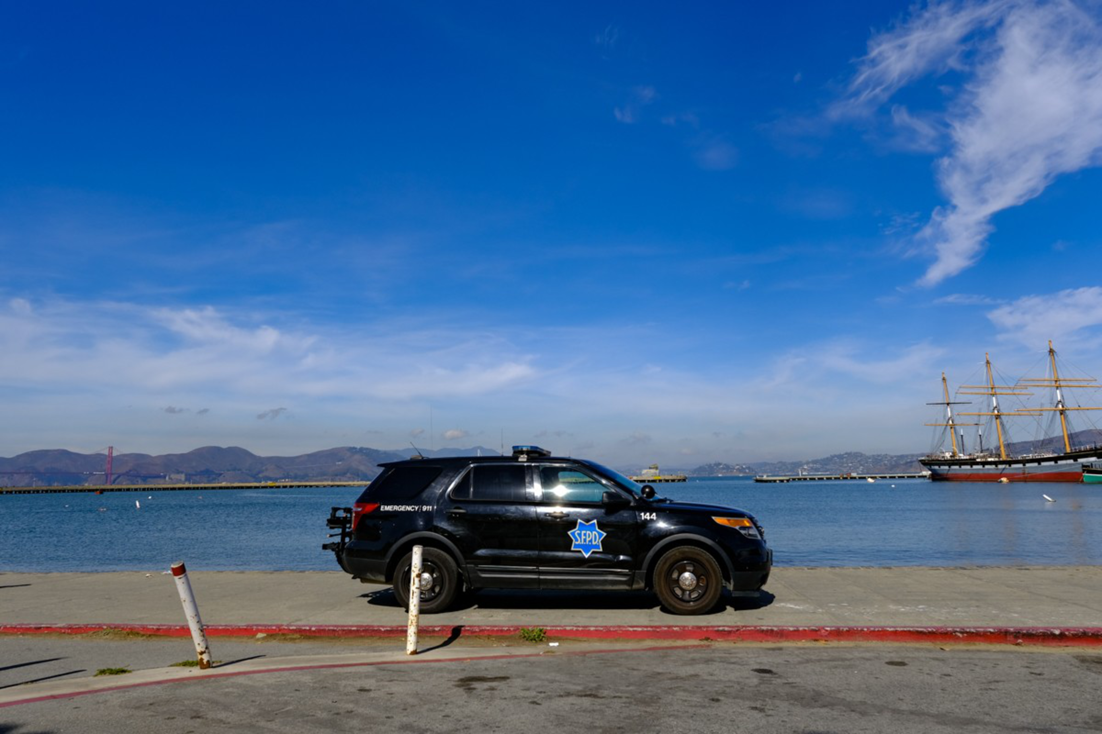 A black police SUV with "SFPD" on the side is parked by a waterfront with calm blue water and distant hills under a bright blue sky.