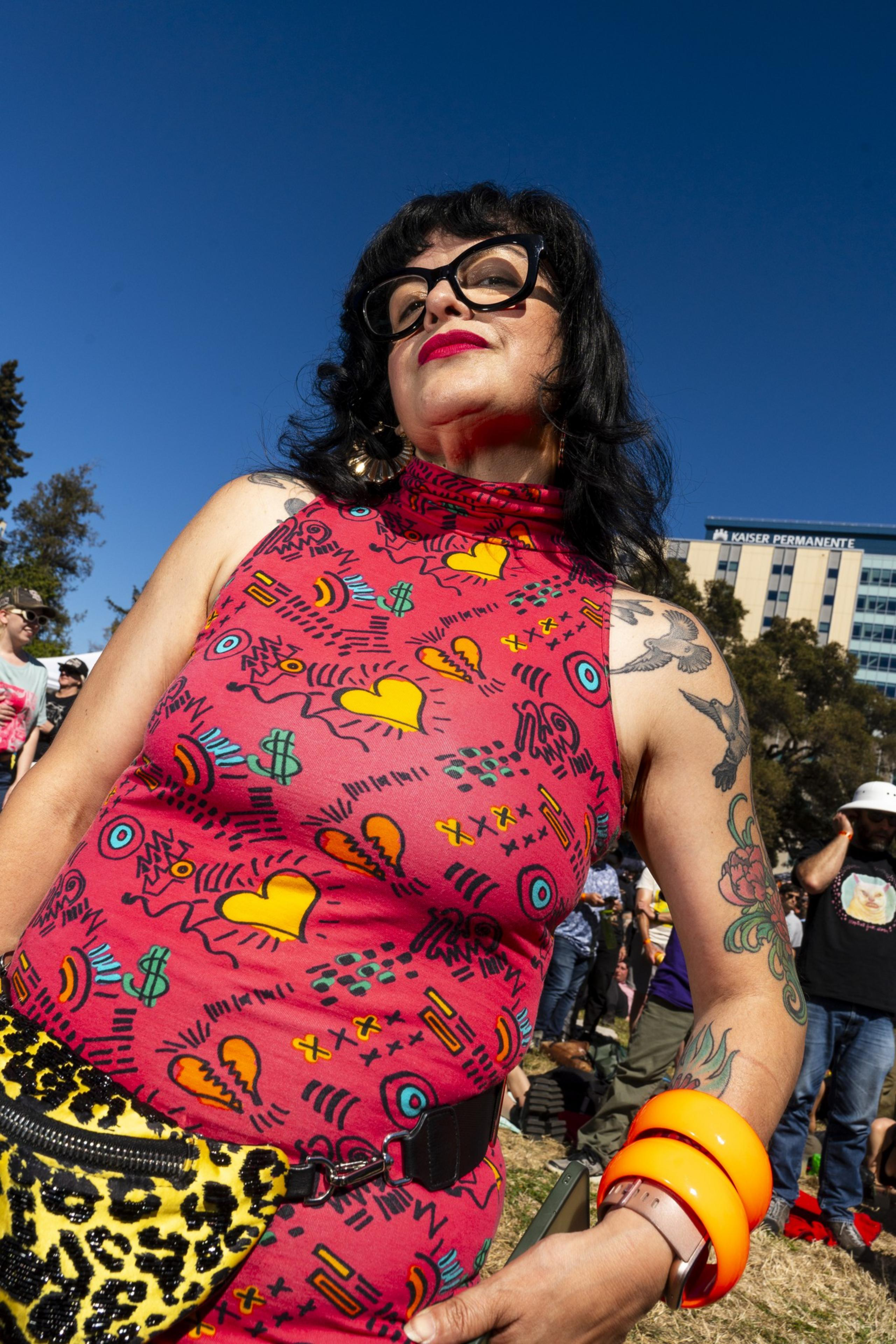 A person with glasses and long dark hair is wearing a colorful, patterned sleeveless top and orange bracelets, standing outdoors under a clear blue sky.