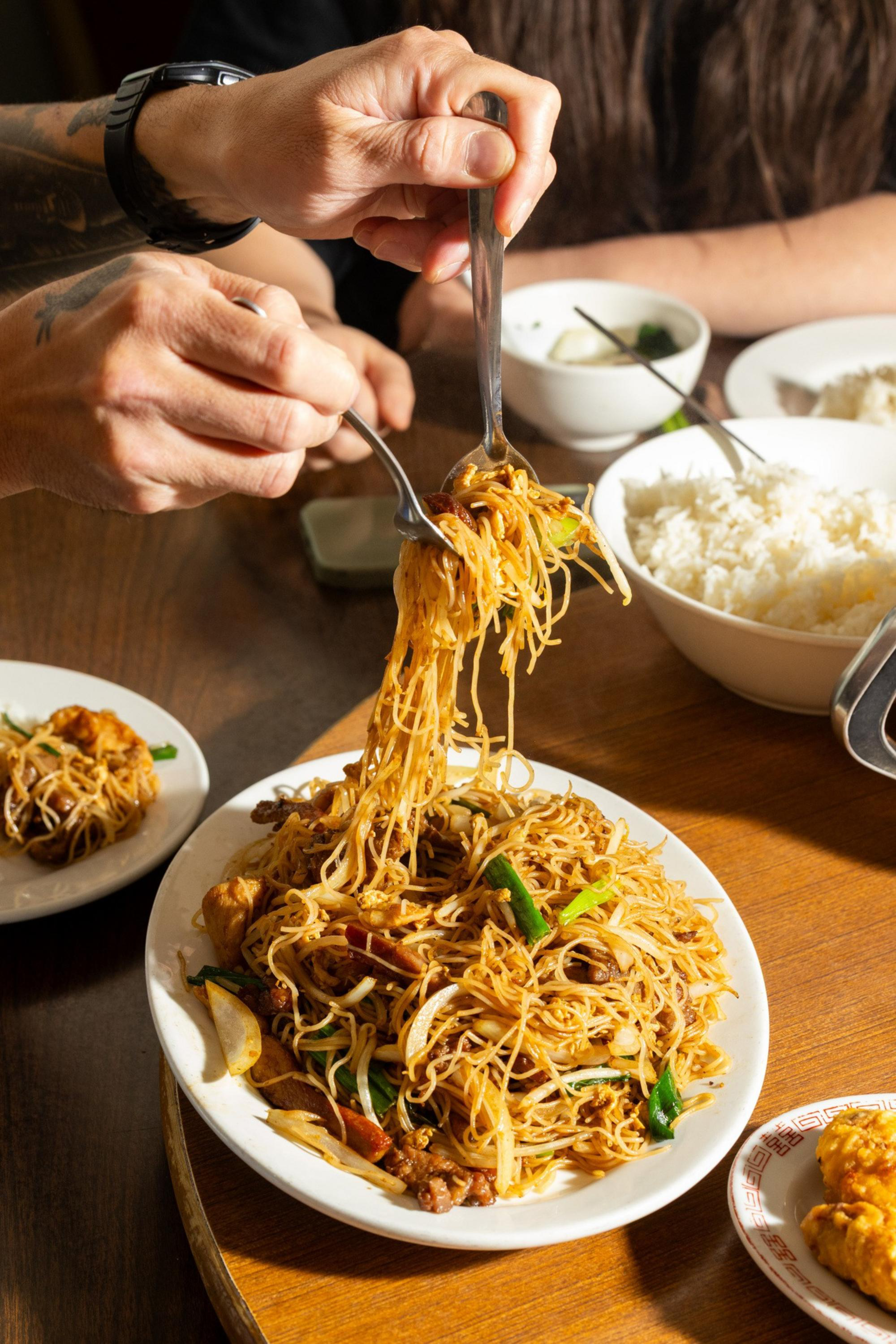 Two hands use fork and spoon to lift stir-fried noodles with vegetables and meat from a plate, with bowls of rice and other dishes on the wooden table.