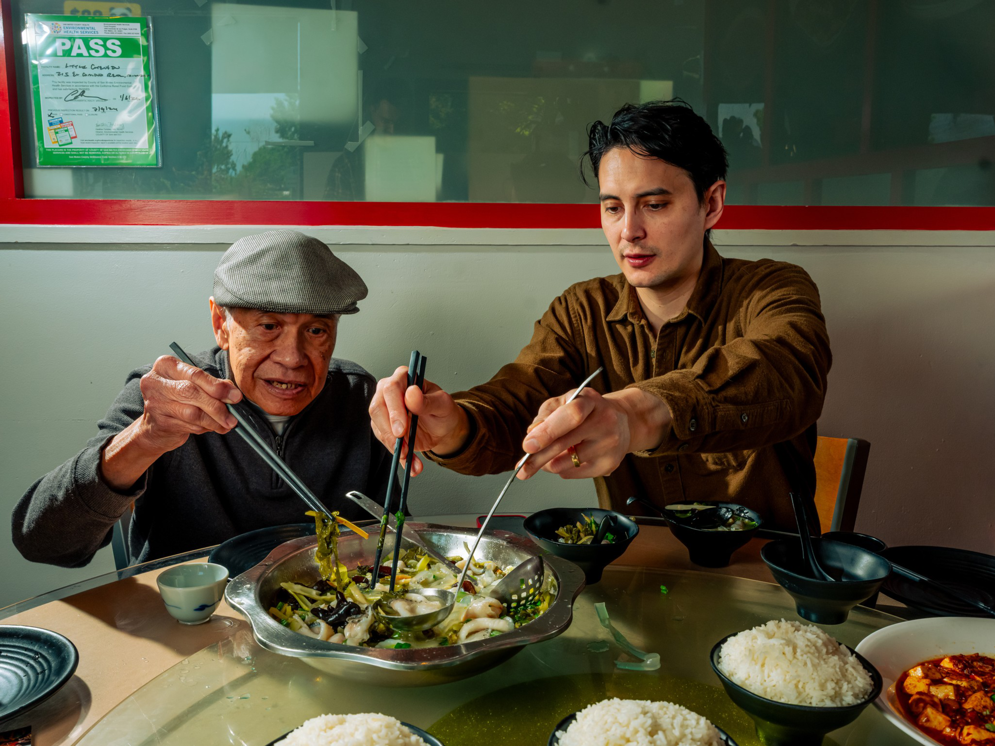 An older man and a younger man use chopsticks to share a meal from a large hot pot on a dining table with bowls of rice and soup.