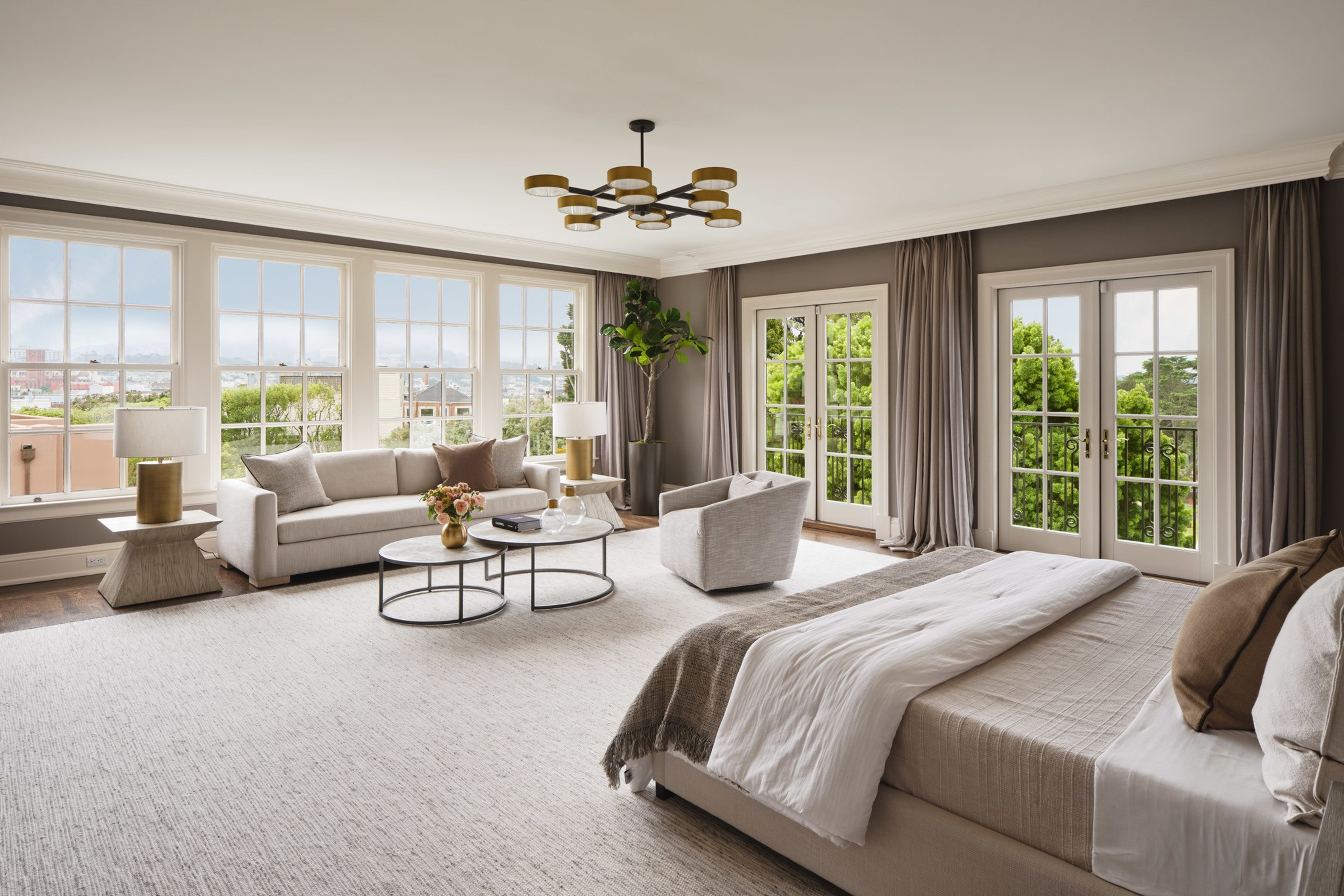 A spacious bedroom with a large bed, a sitting area with a sofa and armchair, large windows, two sets of French doors, and neutral-toned decor.