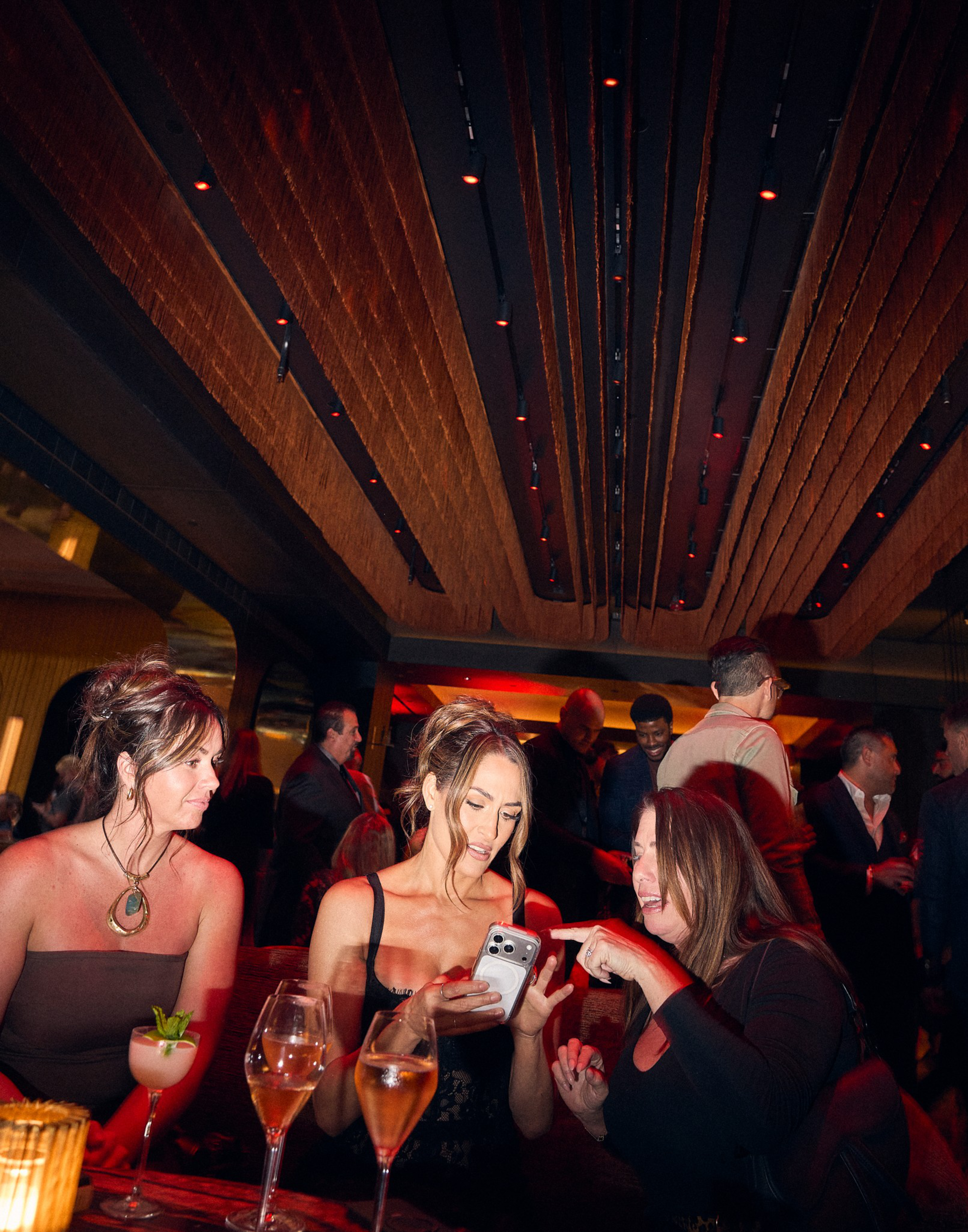 Three women sit at a table with drinks, one showing her phone to another while the third watches, in a dimly lit, crowded, modern lounge.