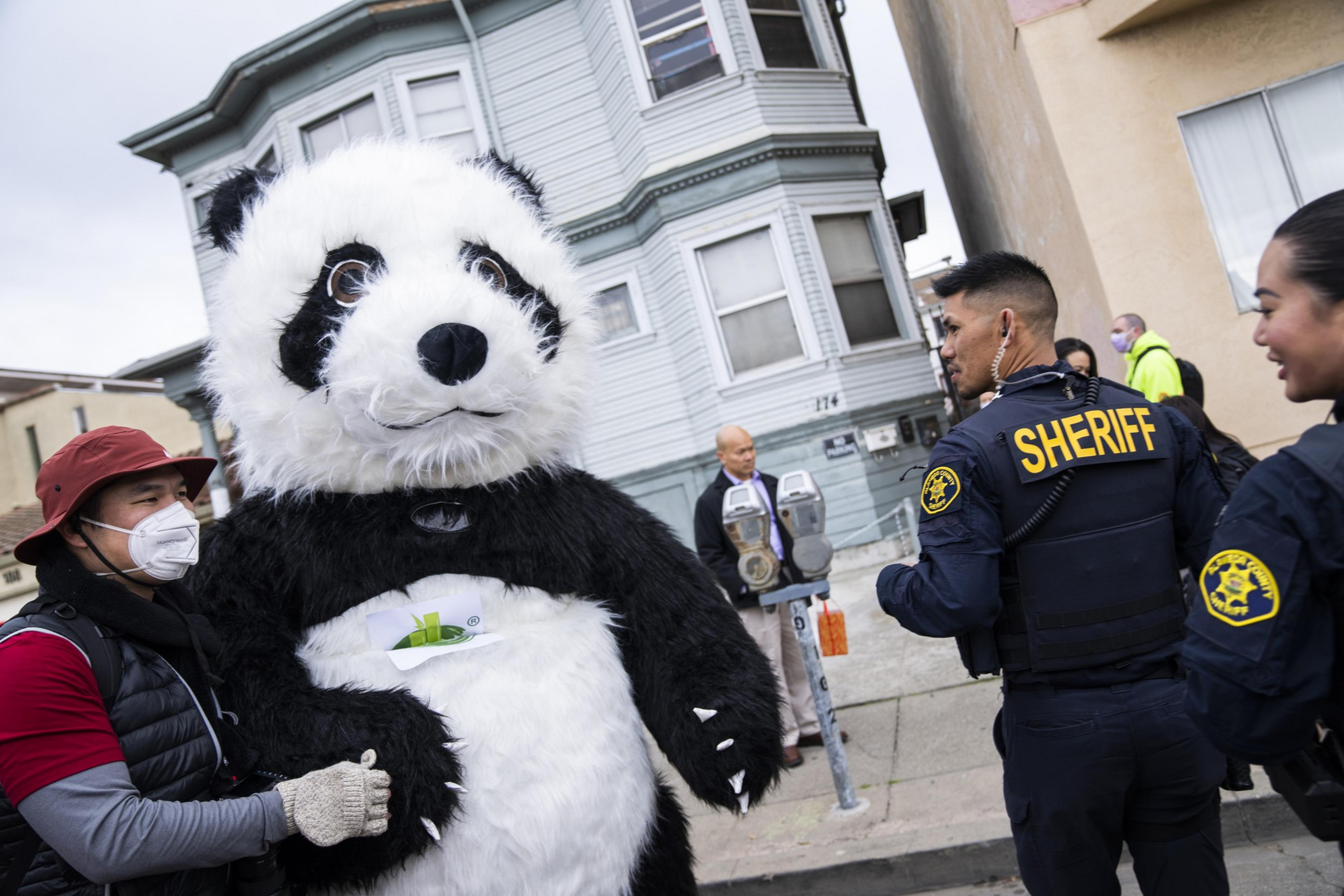A person in a panda costume is being guided by another person wearing a mask and gloves, near two sheriffs and a man on the sidewalk.