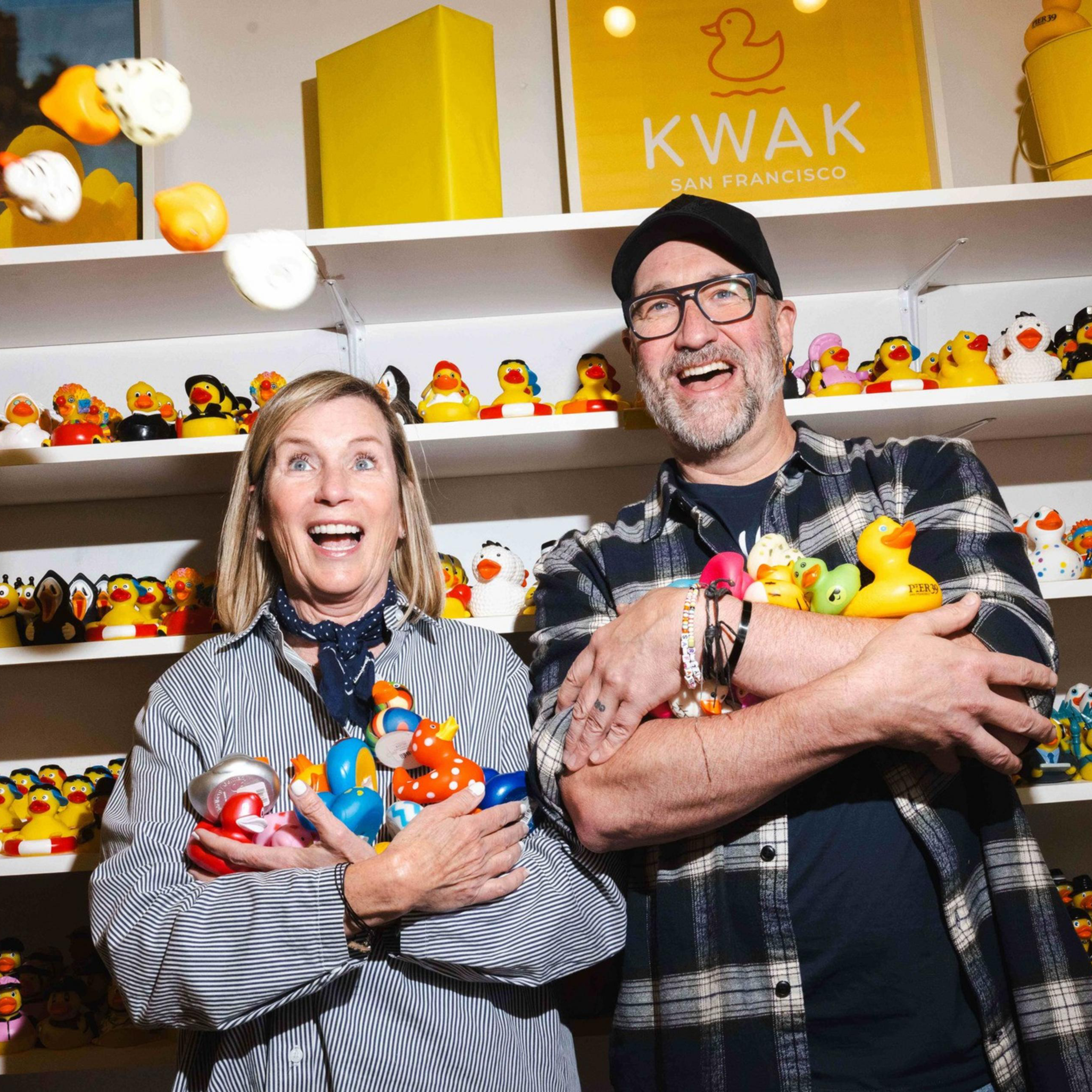 A smiling woman and man stand in front of shelves filled with rubber ducks, each holding several ducks in their arms. Bright lighting highlights their joyful expressions.