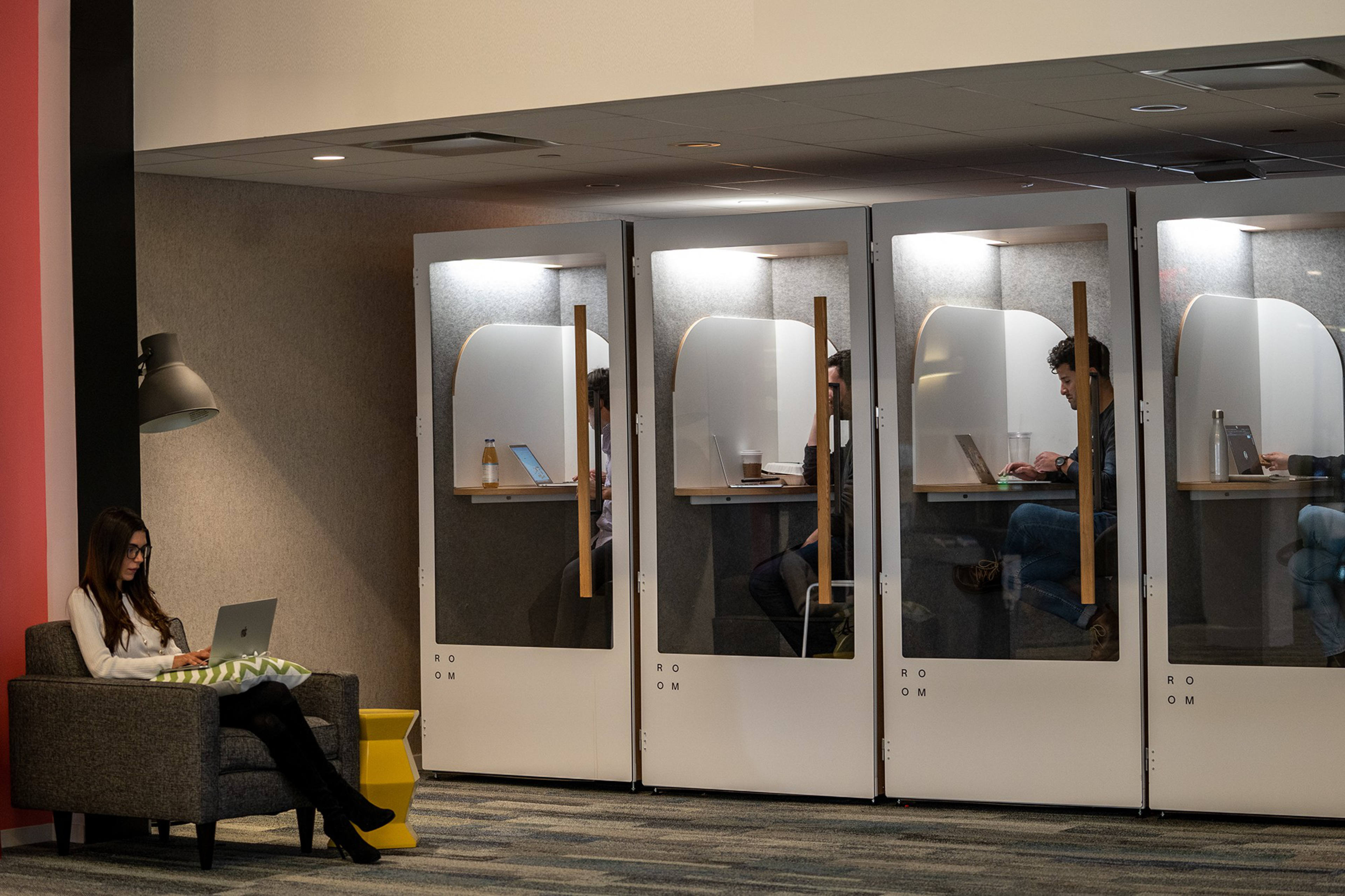 A person sits on a gray armchair with a laptop, while four individuals work in glass cubicles with laptops, each compartment well-lit and containing a small desk.