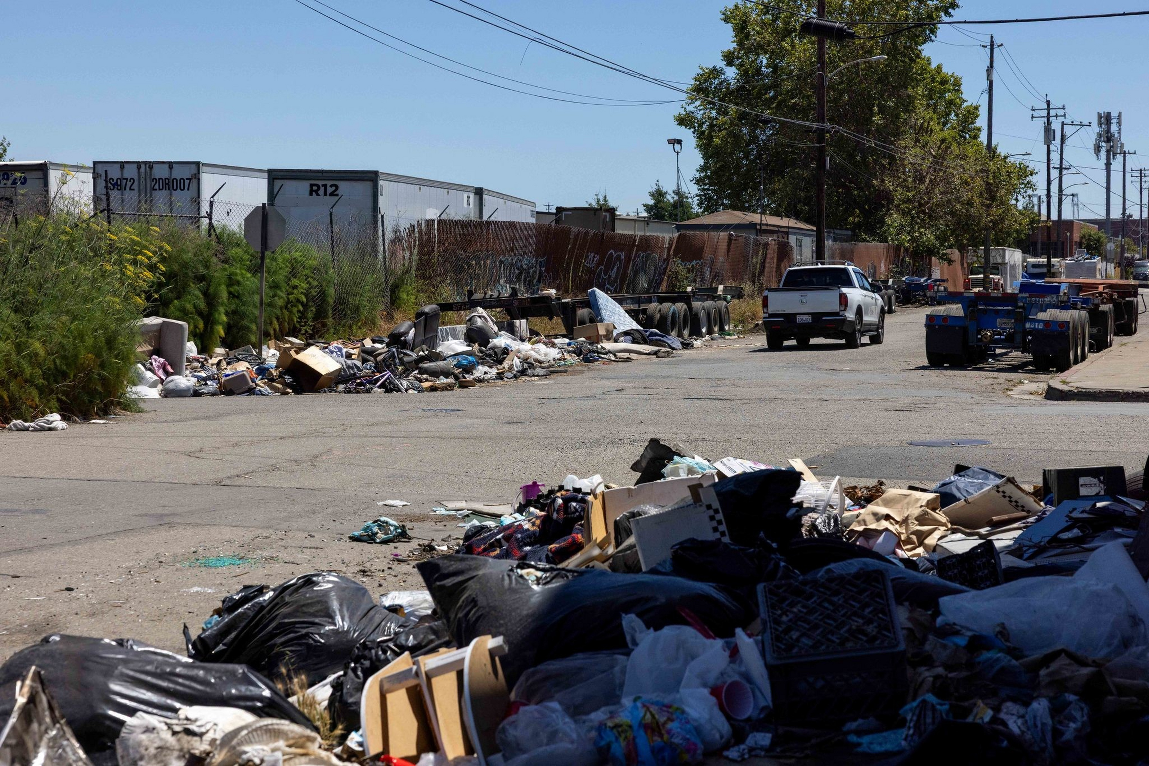 A street filled with piles of trash and debris along the curb, with a white pickup truck and trailers parked further down the road.