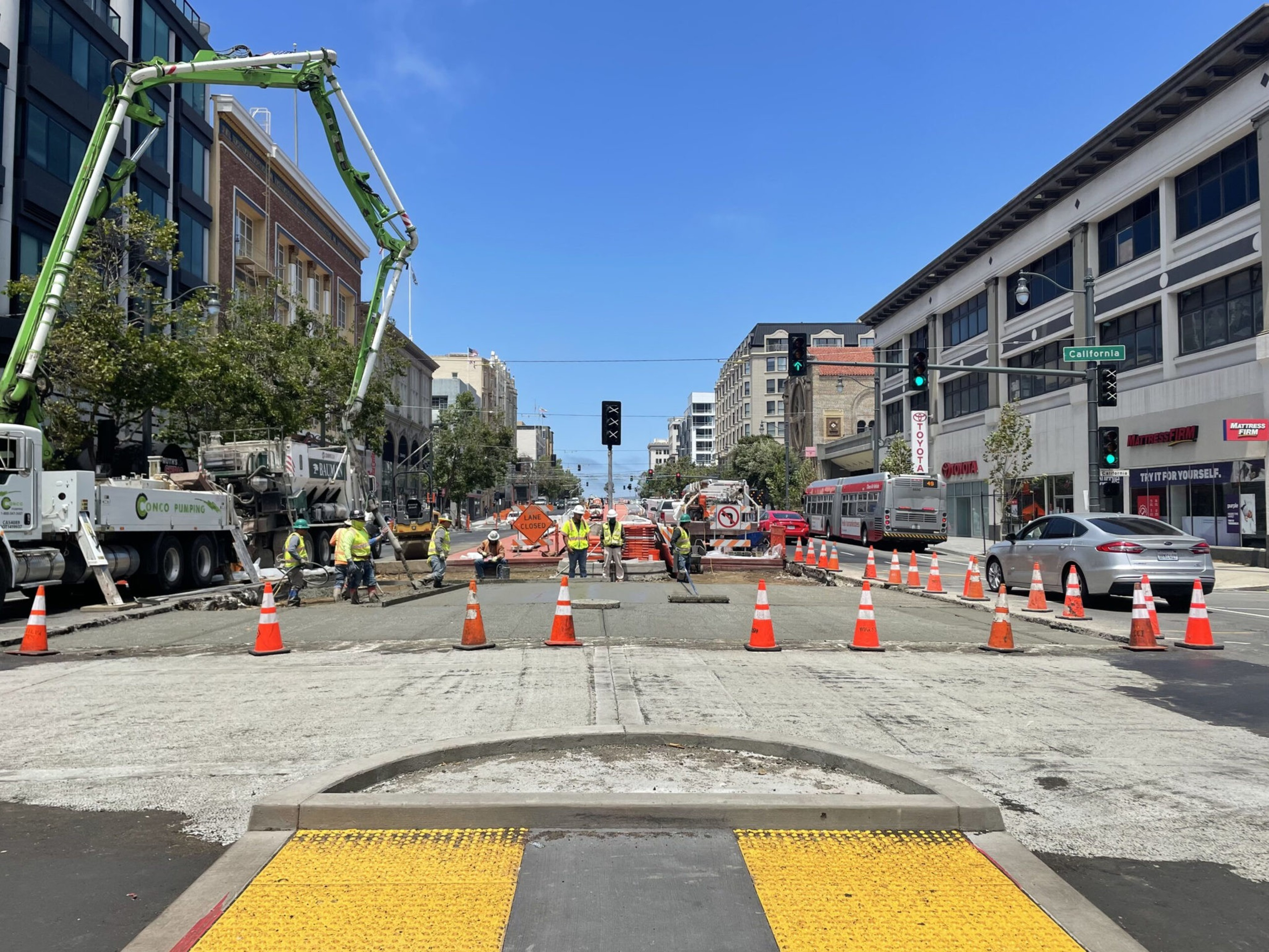 Workers repair a closed city street lined with orange cones, heavy machinery, and traffic signs, with buildings and vehicles flanking the road under a clear blue sky.