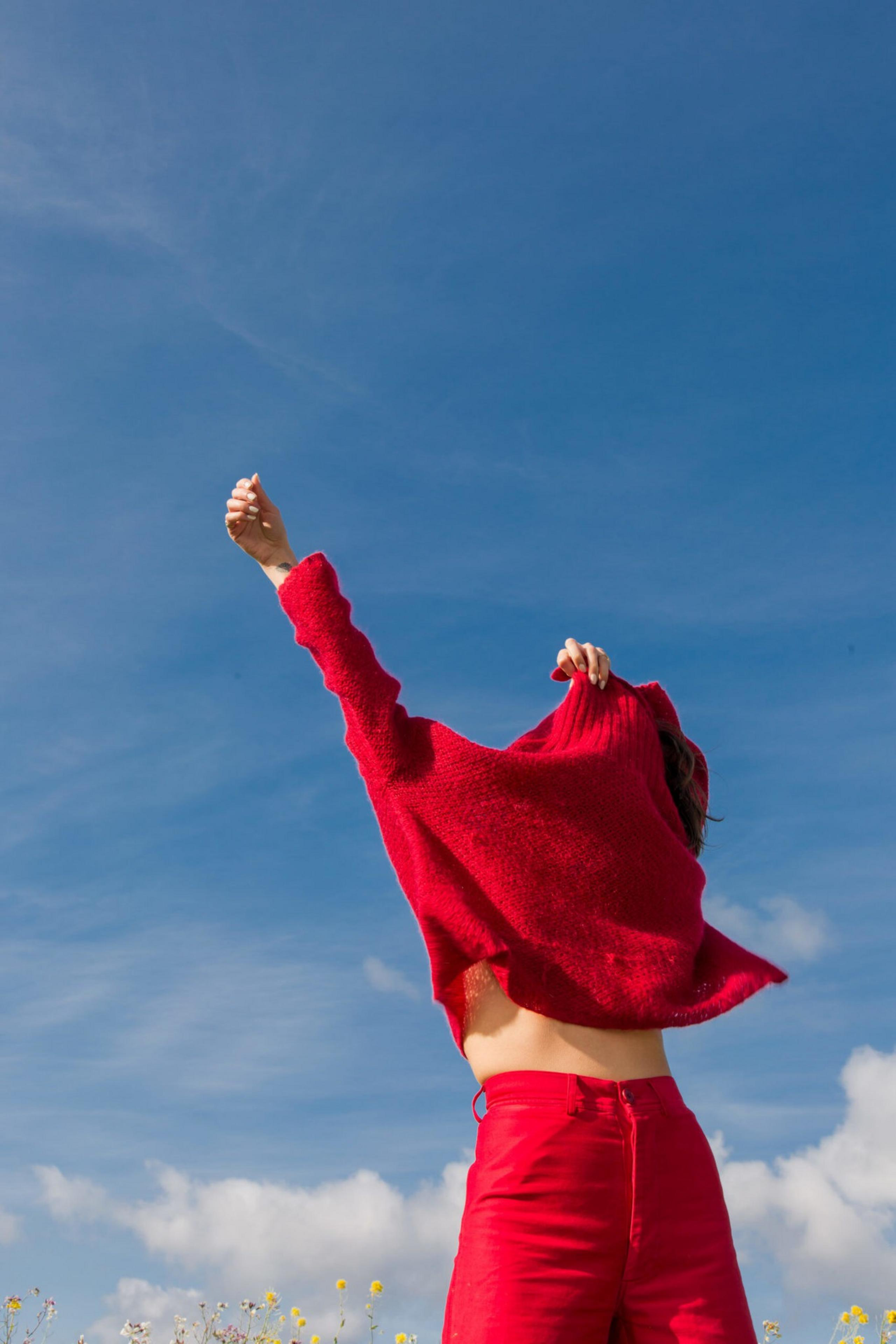 A person wearing bright red pants lifts a matching red sweater over their head, standing against a blue sky with scattered clouds and some wildflowers below.