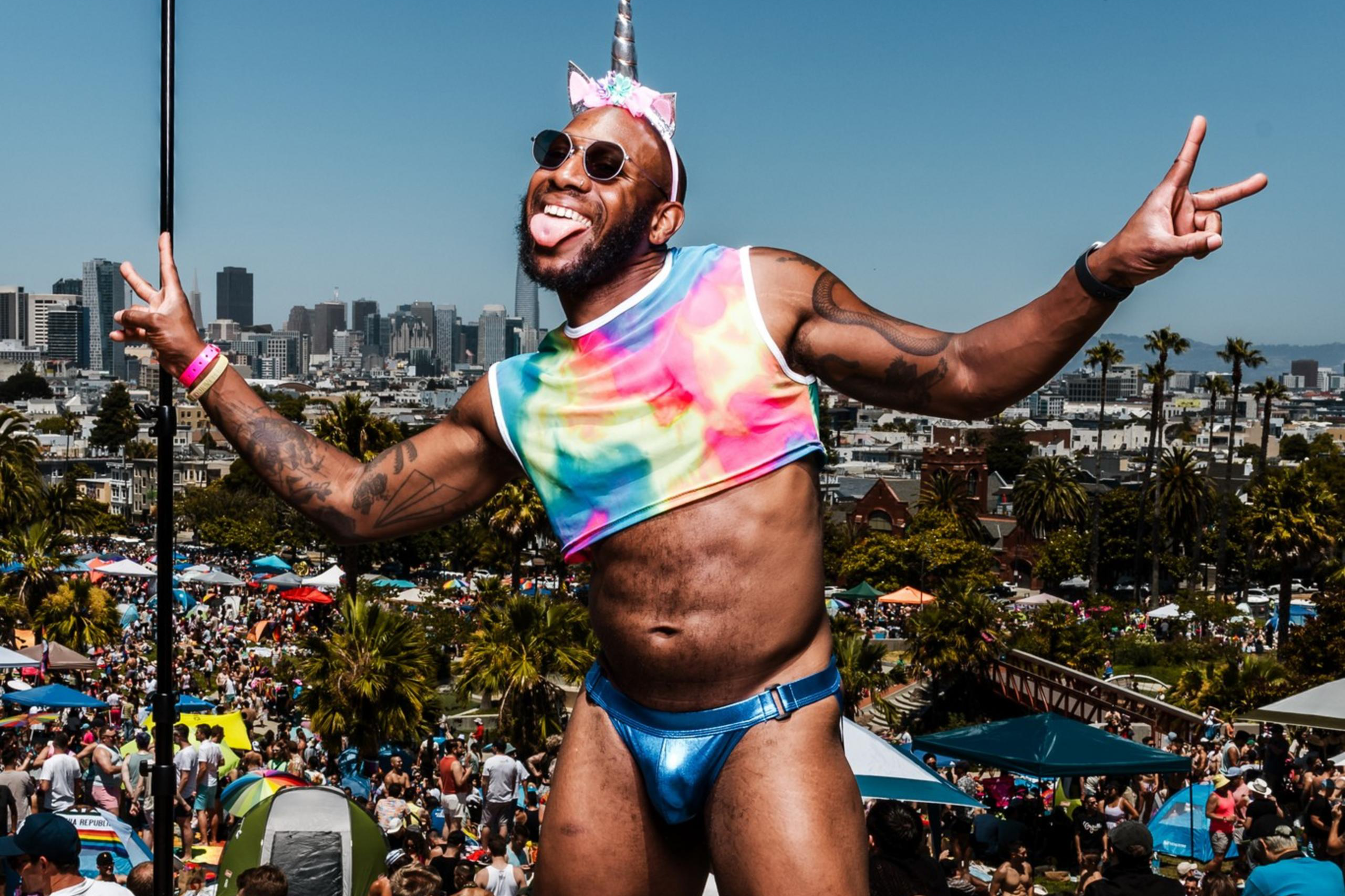 A person dressed in a rainbow crop top, blue briefs, and a unicorn headband with a horn poses joyfully, making a peace sign. They are in a park crowded with people.