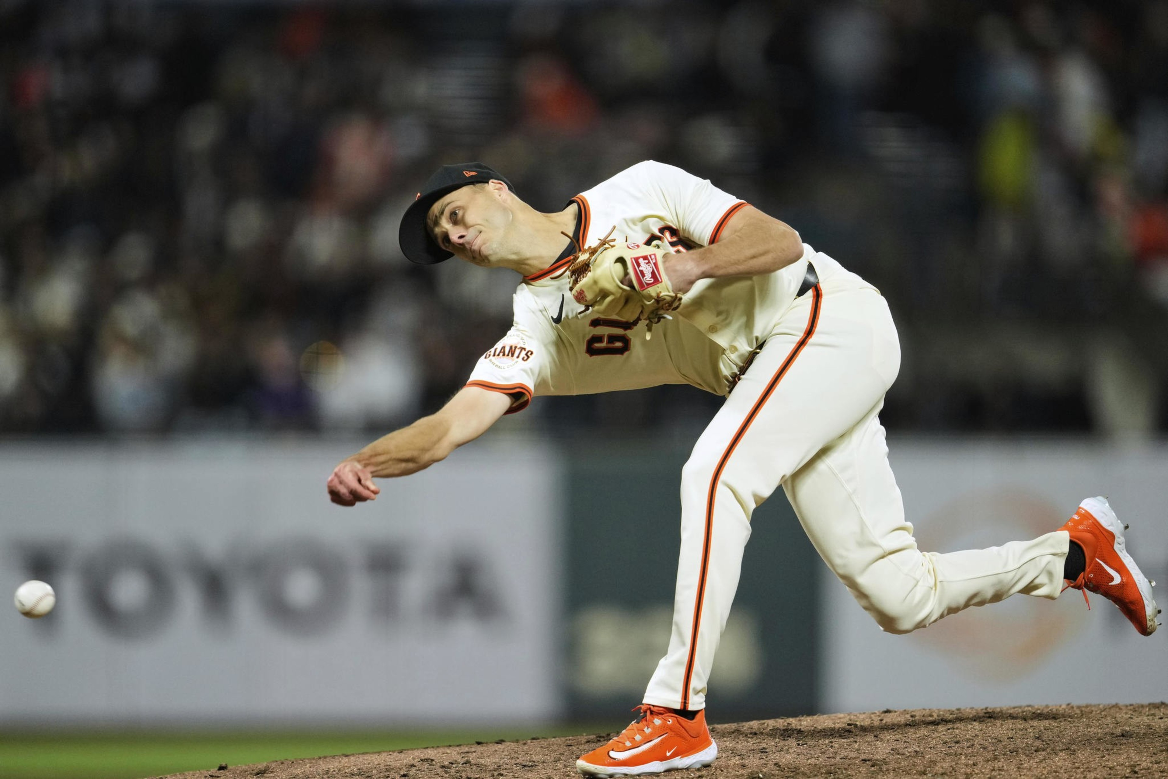A baseball player wearing a white uniform and orange shoes pitches the ball mid-action. The background is a blurred stadium.
