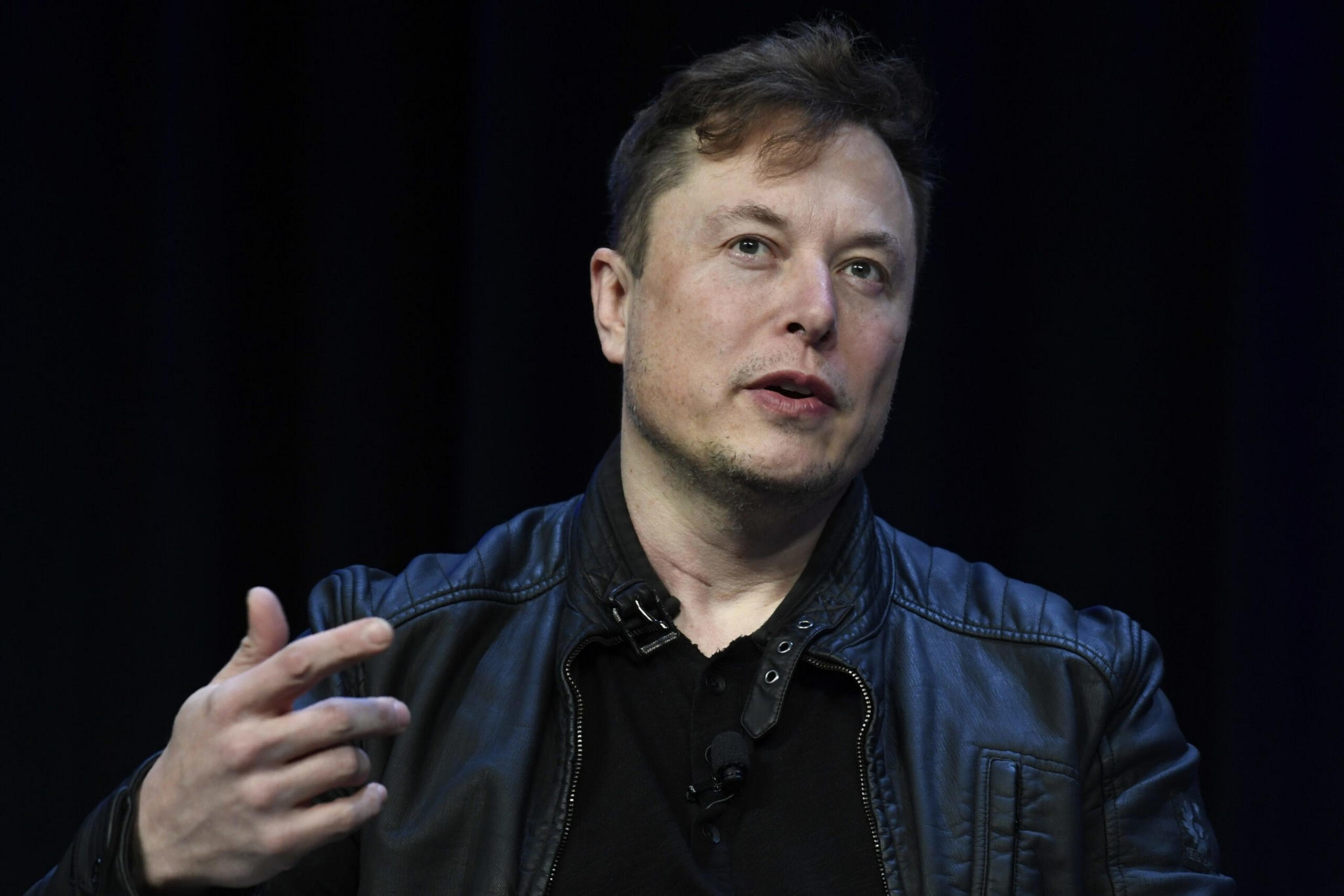 A man with light skin and short brown hair wears a black leather jacket and black shirt, gesturing with his right hand while speaking.