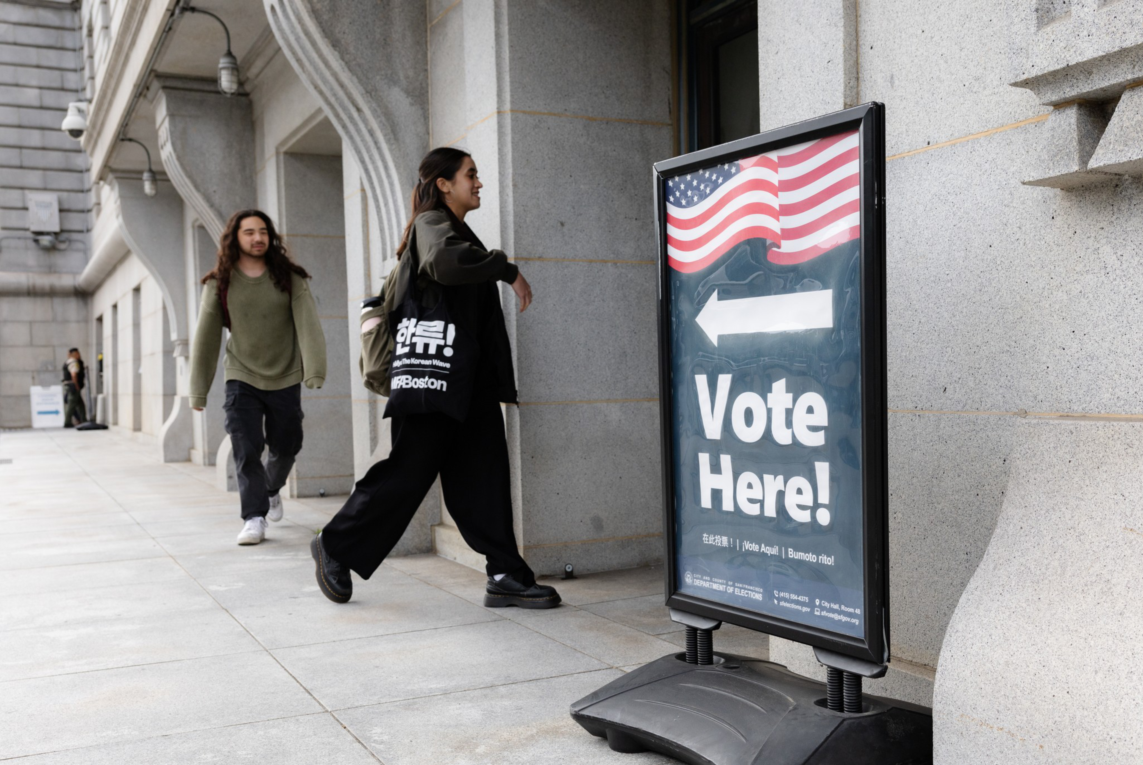 A woman approaches a "Vote Here!" sign with an American flag graphic, while a man walks behind her on a city sidewalk near a stone building.