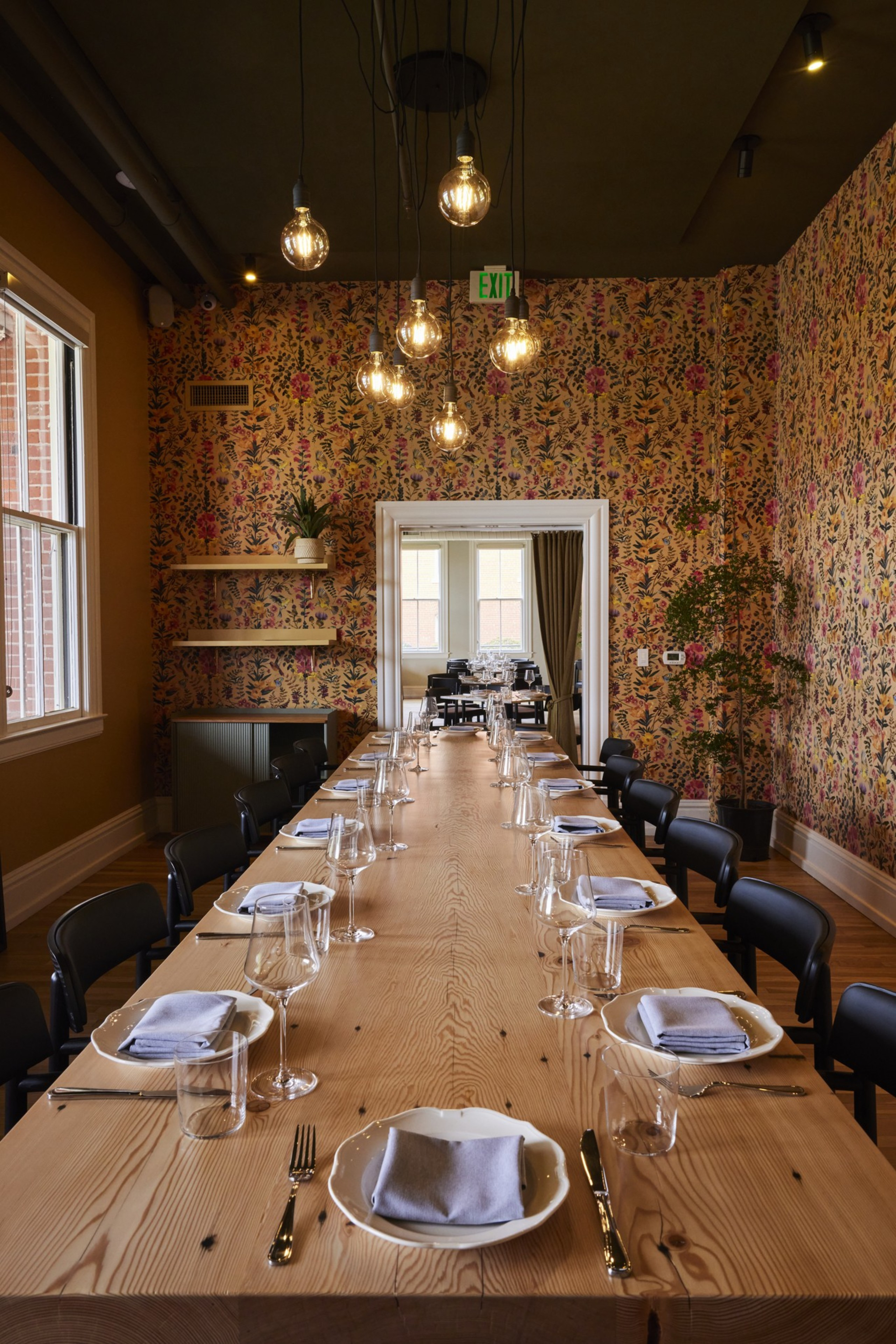 A long wooden table is set with plates, glasses, and napkins in a room with floral wallpaper and hanging bulbs. There is a window and a plant on the side.