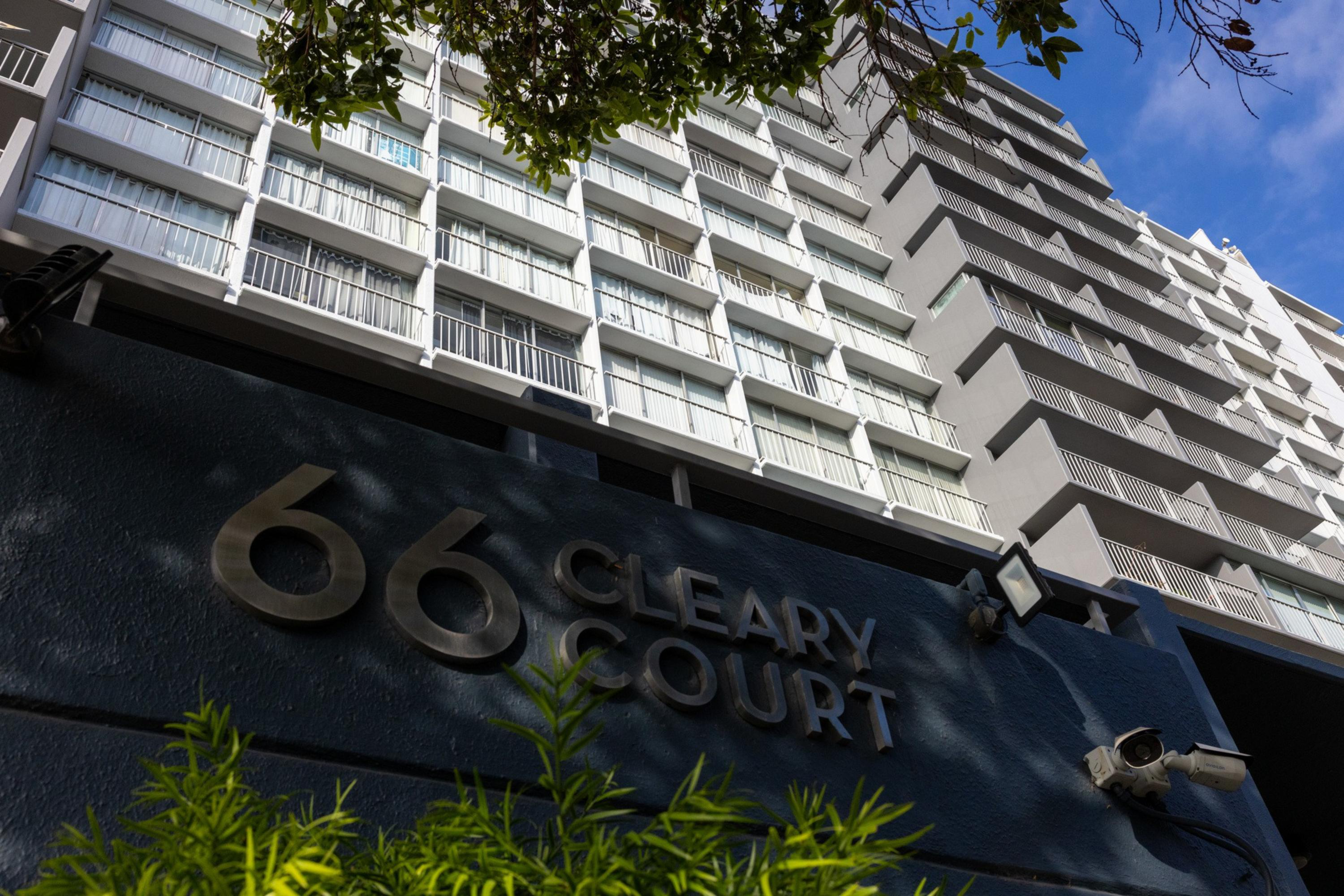 A multi-story white apartment building with balconies stands behind a dark wall displaying “66 Cleary Court” in metallic letters, with plants and security cameras visible.