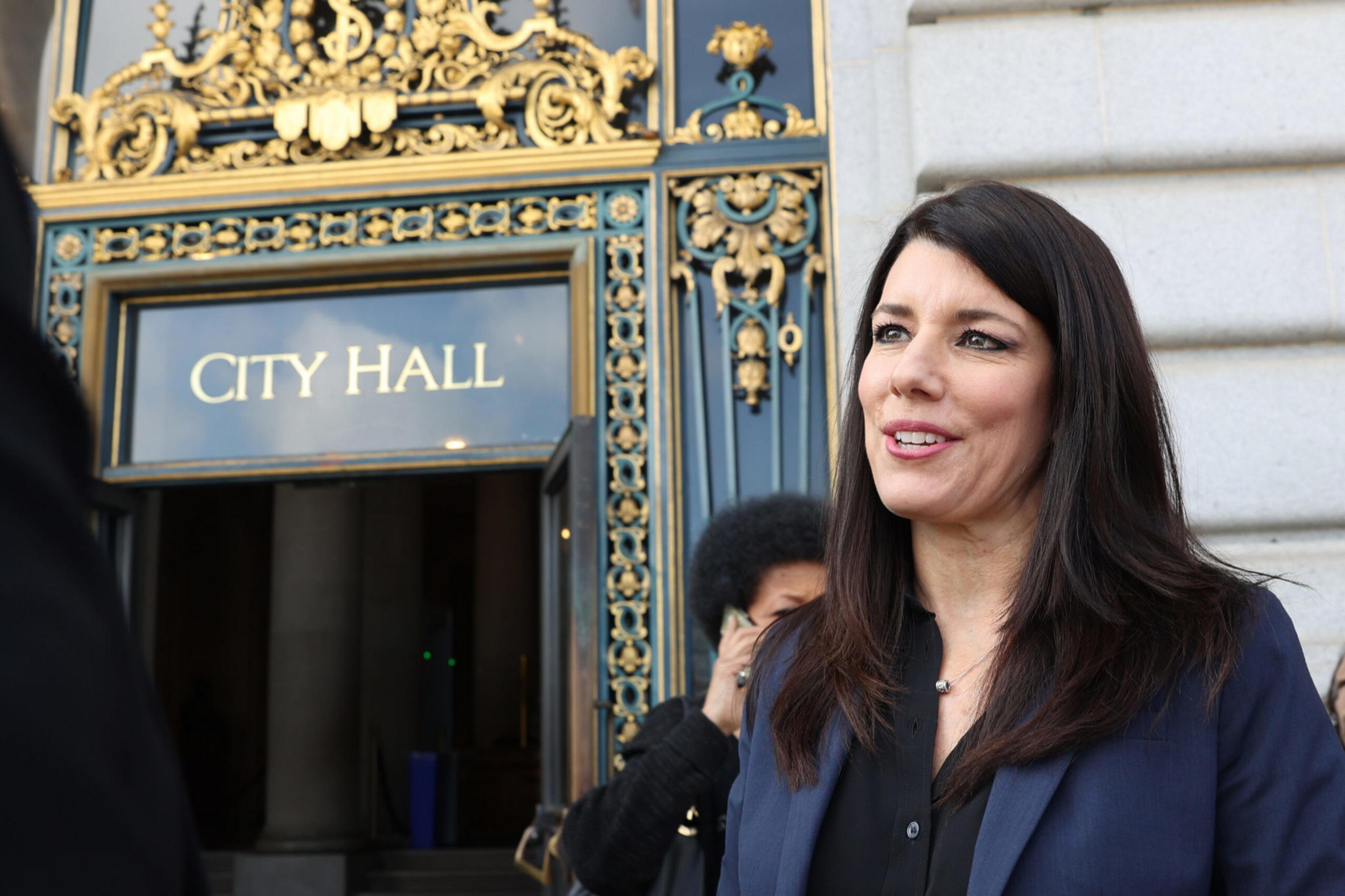A woman with long dark hair wearing a blue blazer stands outside an ornate City Hall entrance with gold and black decorative details.